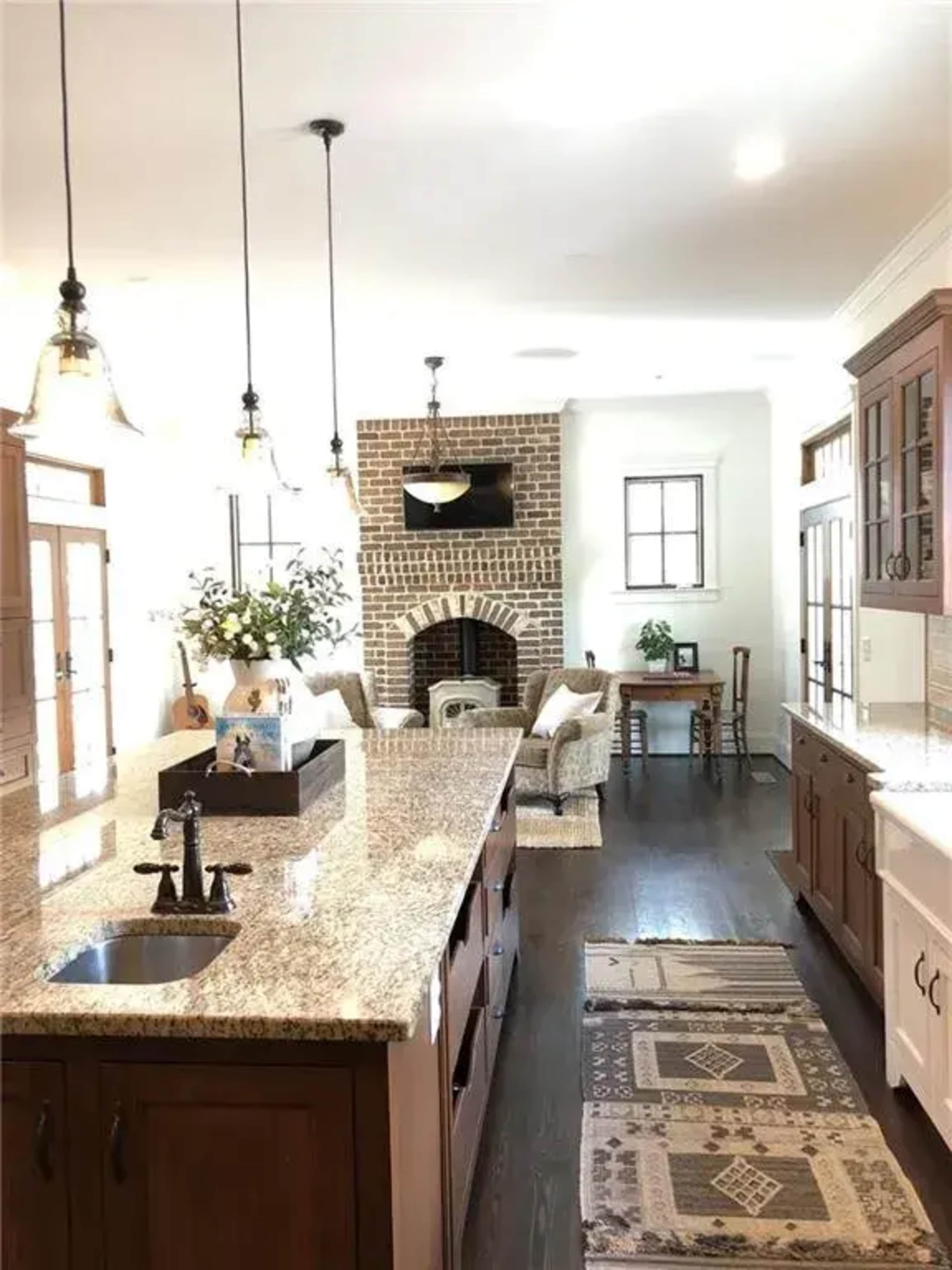 A spacious kitchen features a central island with a granite countertop, wooden cabinetry, and a visible living area with a brick fireplace in the background.