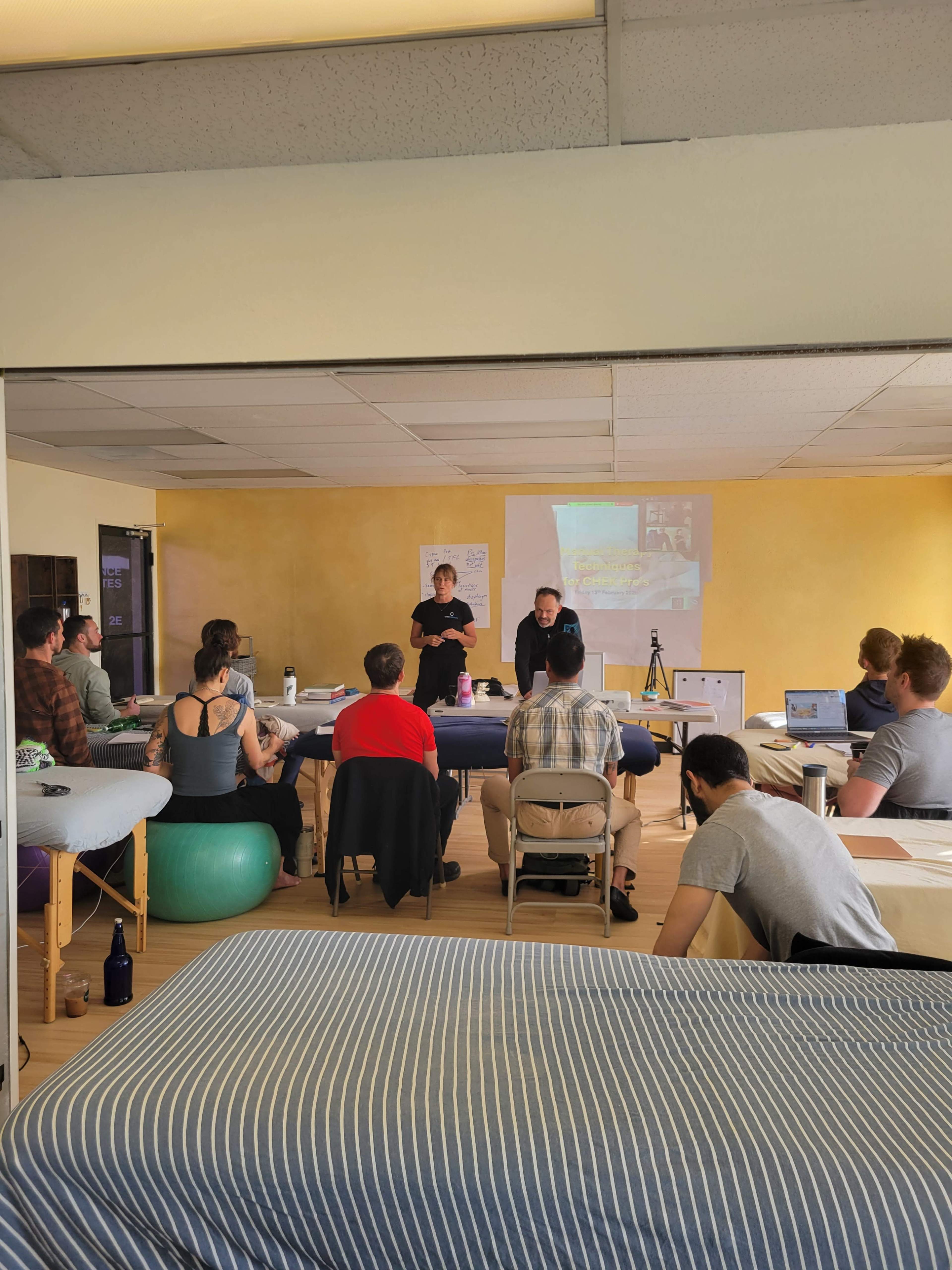 A classroom setting with a group of students seated at tables, listening to two instructors presenting information.