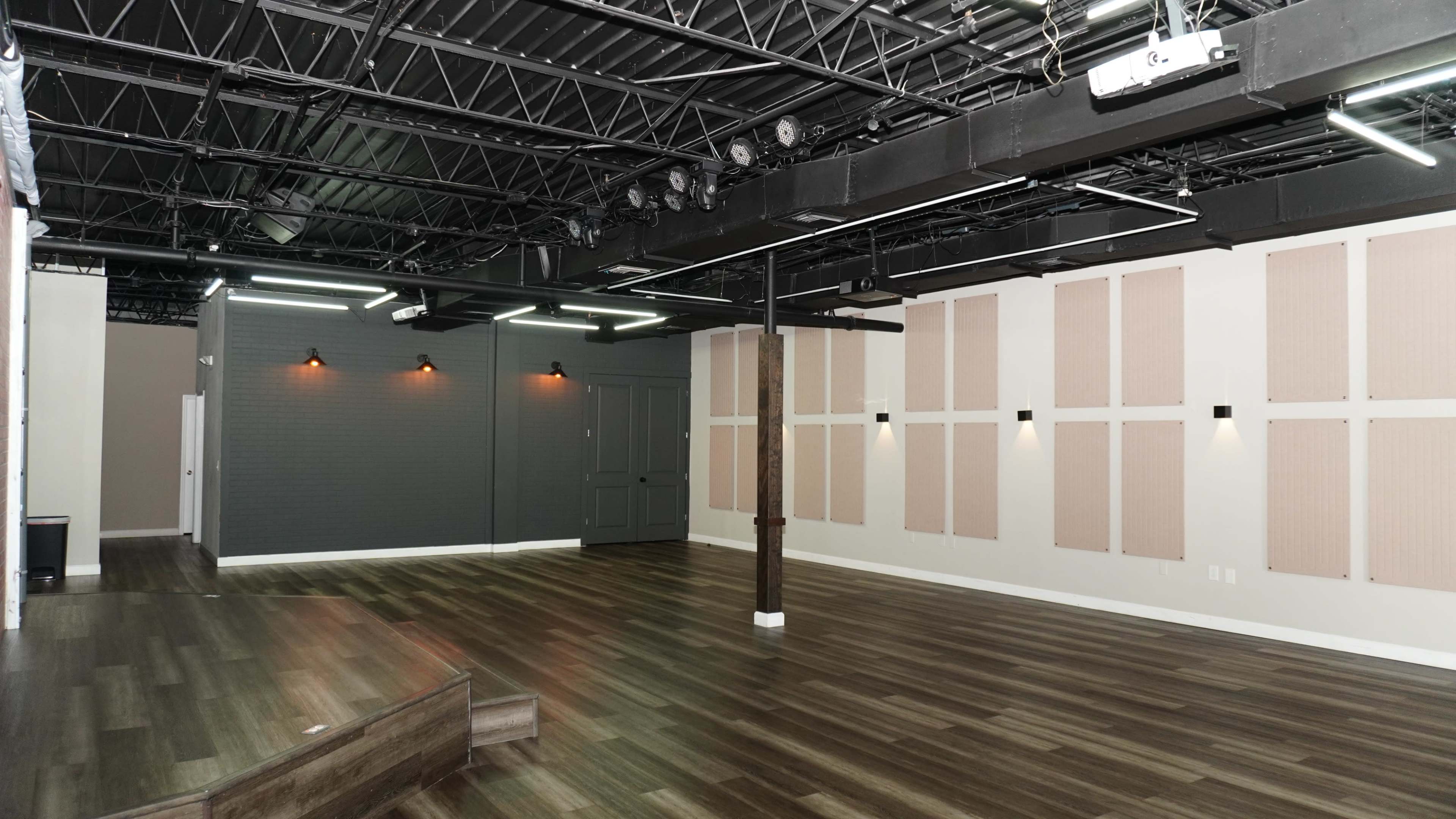 The image shows a spacious, empty room featuring dark hardwood flooring, a gray wall, and acoustic panels arranged on the white walls.