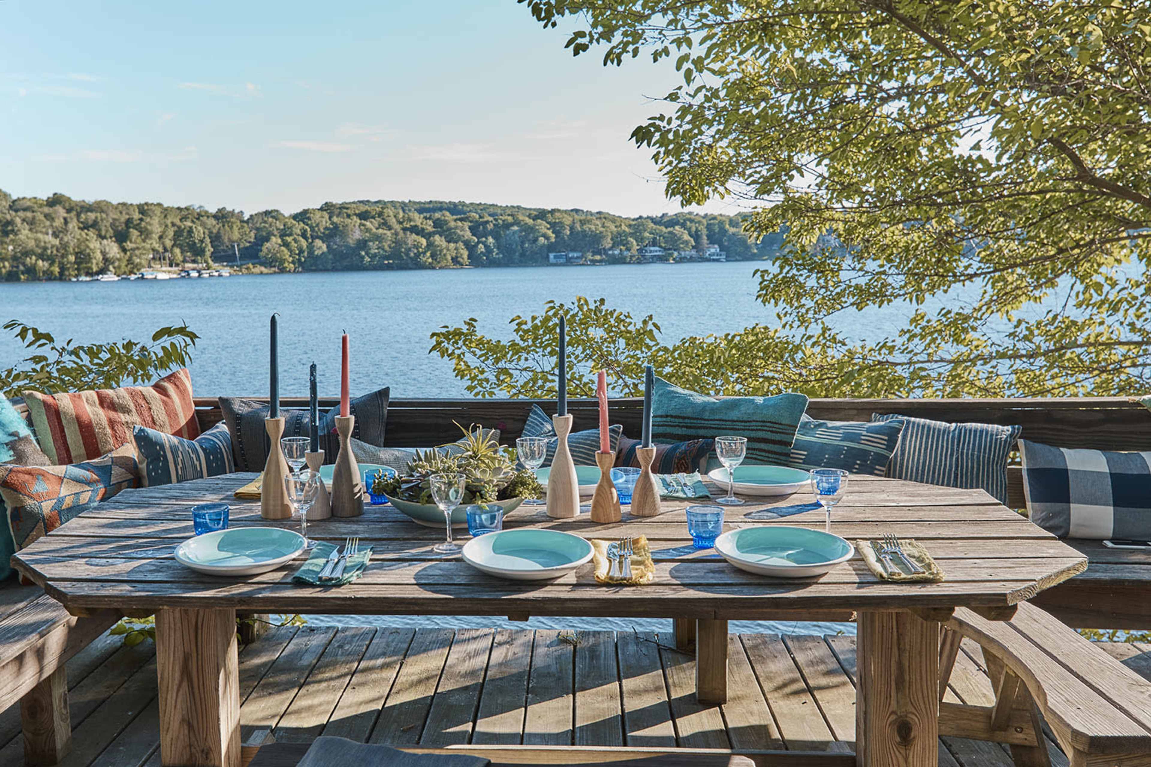 A wooden dining table is set with colorful plates and glassware, overlooking a serene lake surrounded by greenery.