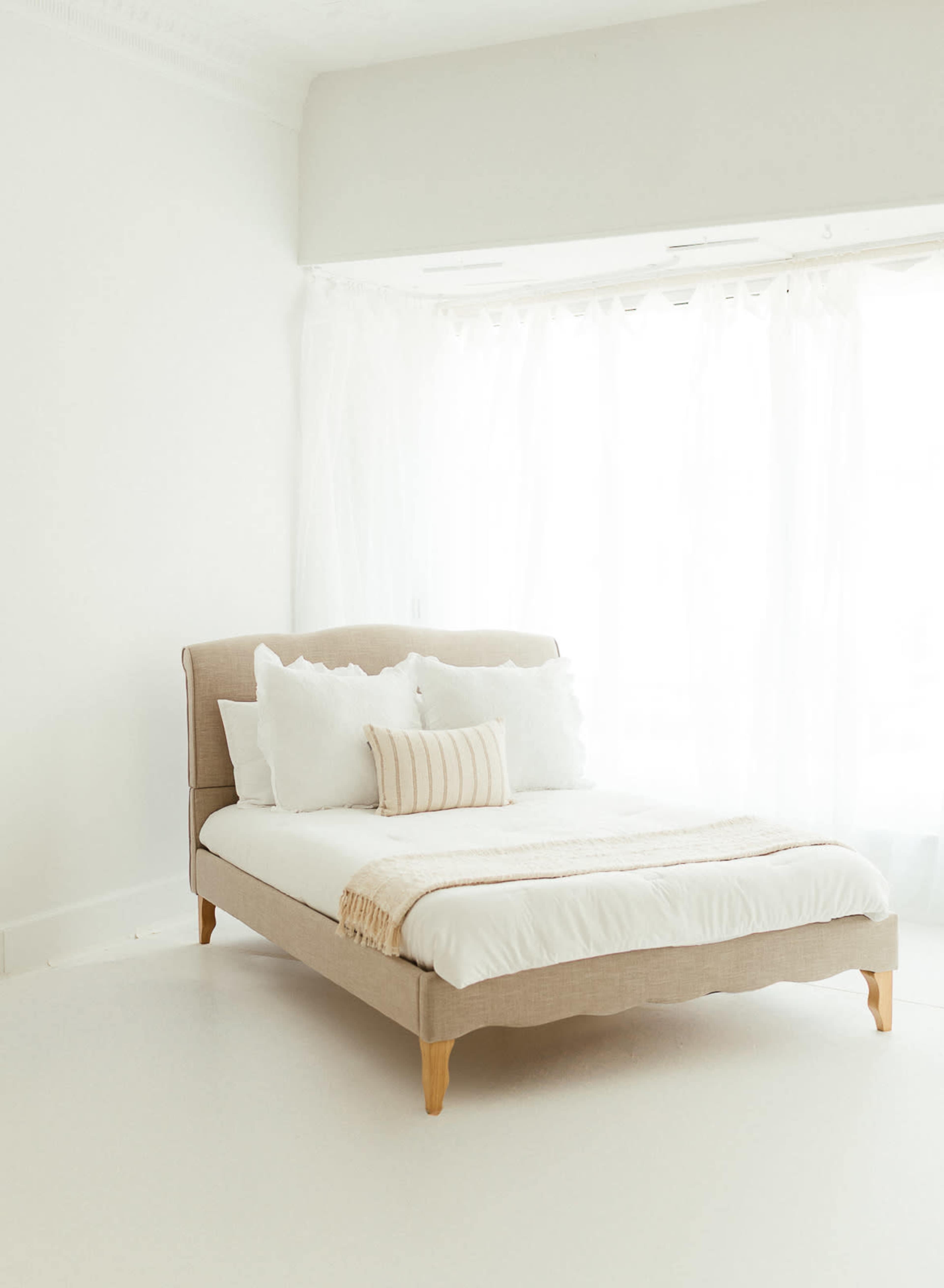 A light-filled bedroom features a bed with a neutral fabric headboard, white bedding, and decorative pillows against a backdrop of sheer curtains.