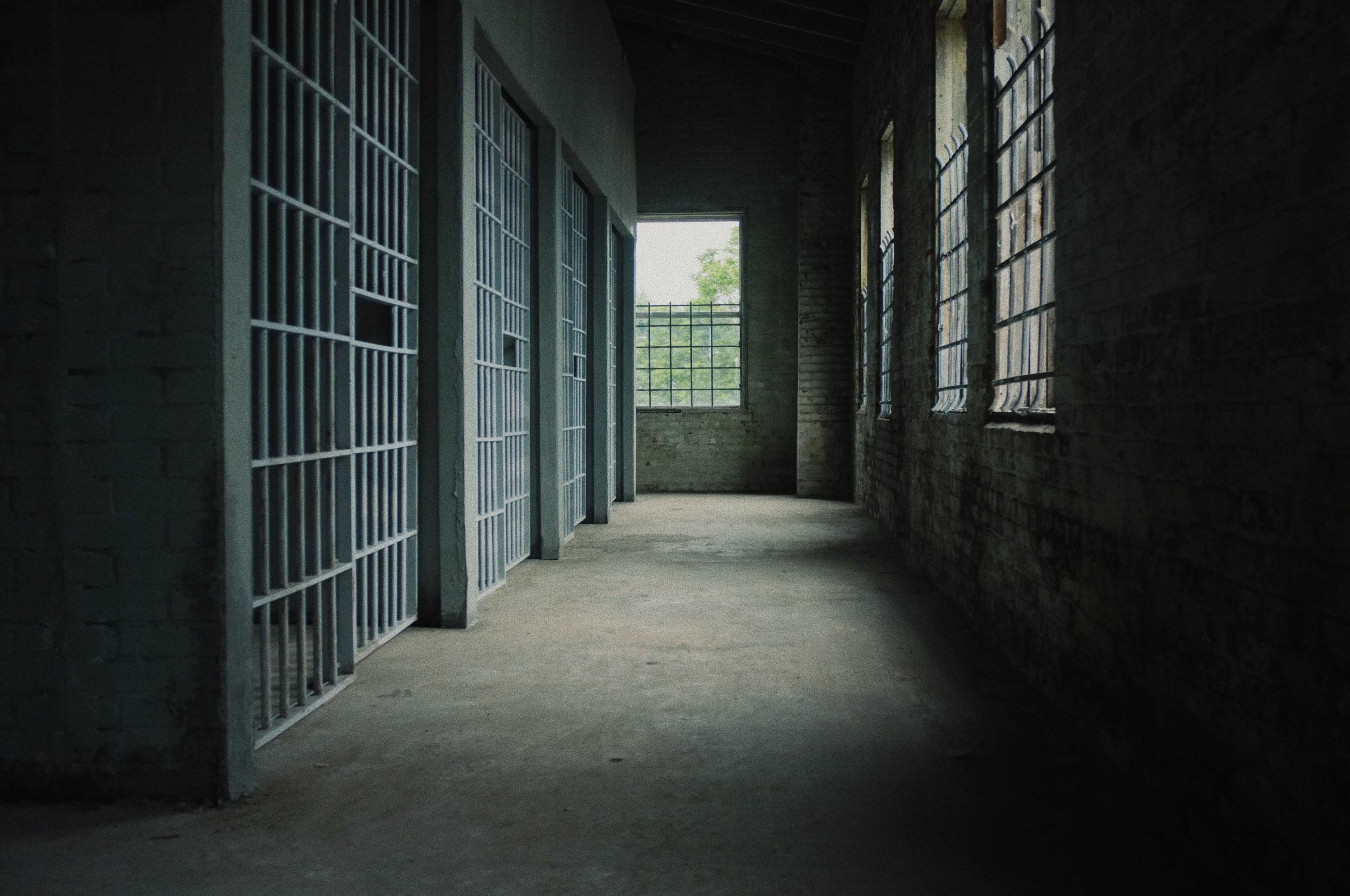 The image shows a dimly lit hallway in an old building with empty jail cells on either side and windows allowing natural light to enter.