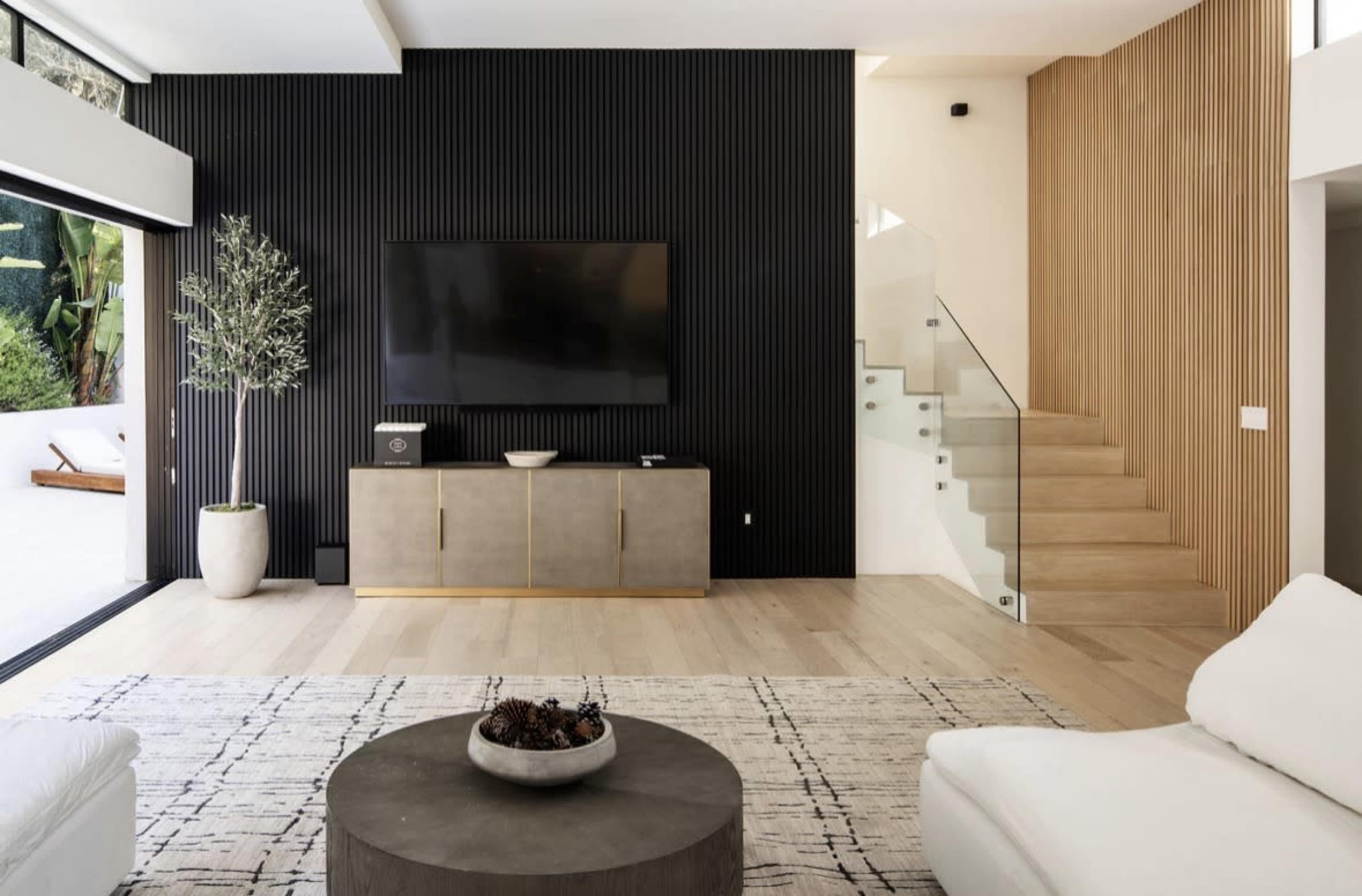 The image shows a modern living room featuring a large television mounted on a black accent wall, a wooden cabinet, a round coffee table in the center, and a staircase with wooden paneling.