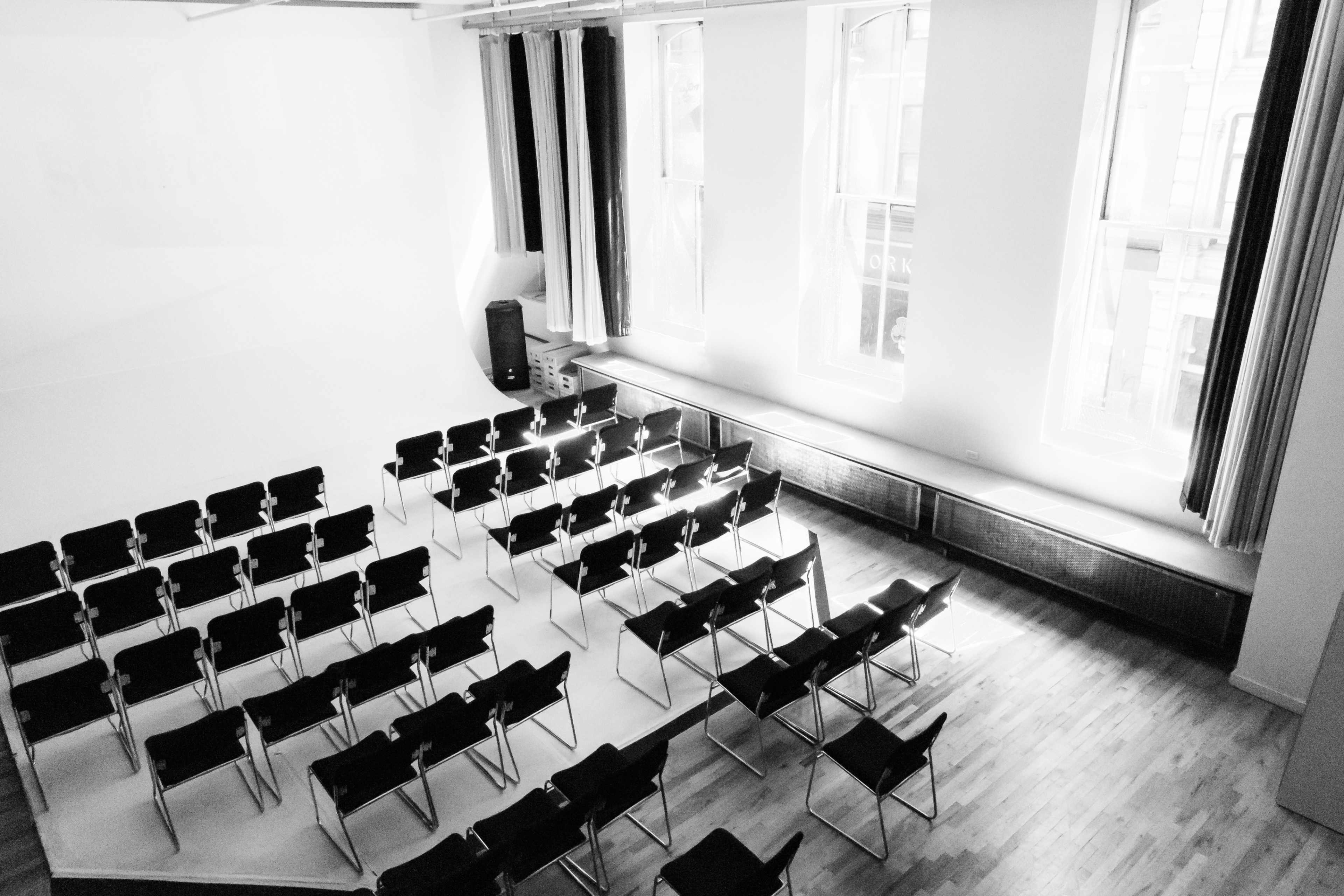 A row of black chairs is arranged in a semi-circle facing a blank wall in a well-lit room with large windows.