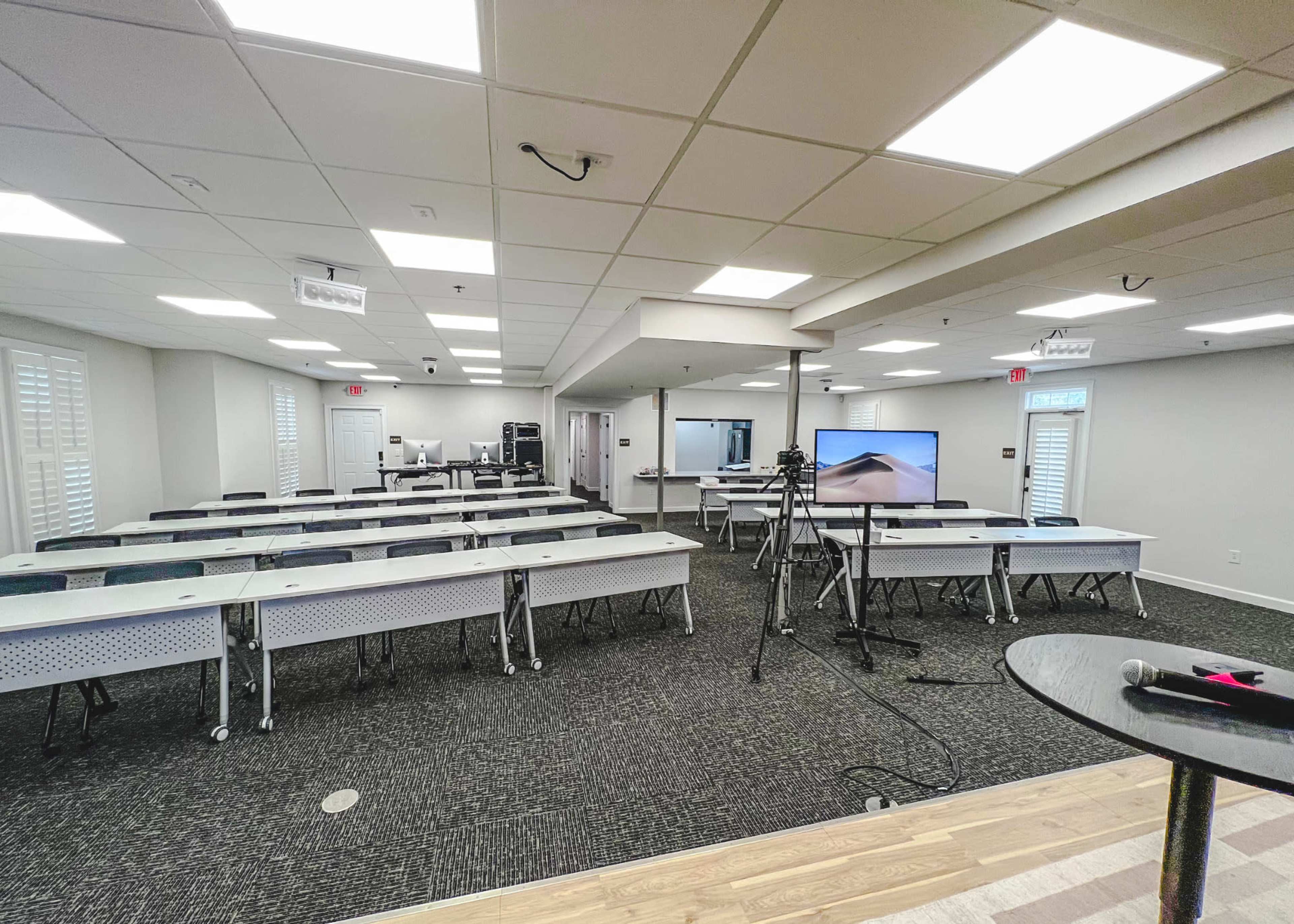 A well-lit conference room set up with rows of tables and chairs, an empty podium, and a large screen displaying a landscape.