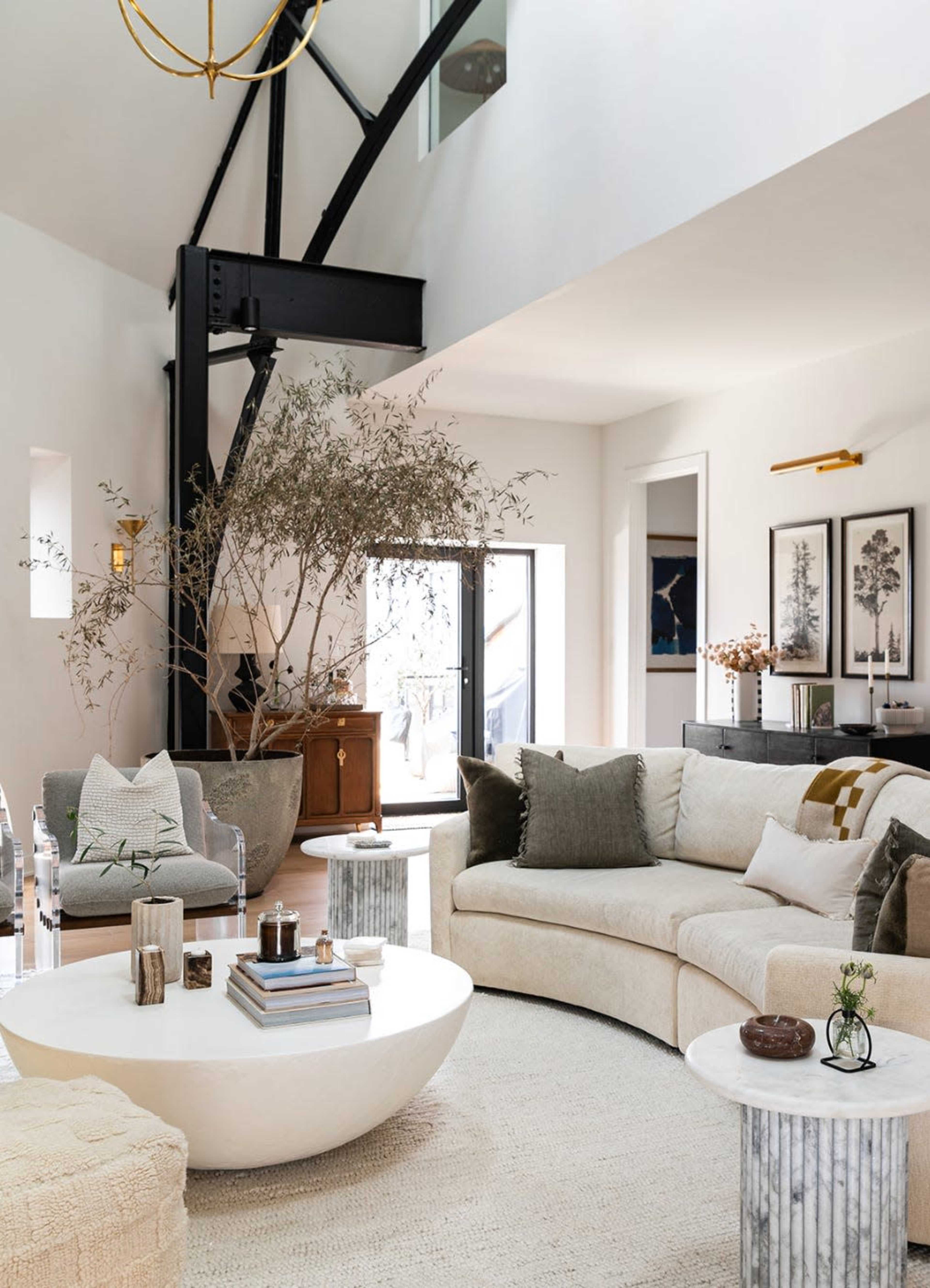 A modern living room features a curved sectional sofa, a round coffee table, and a tall indoor plant, with large windows letting in natural light.