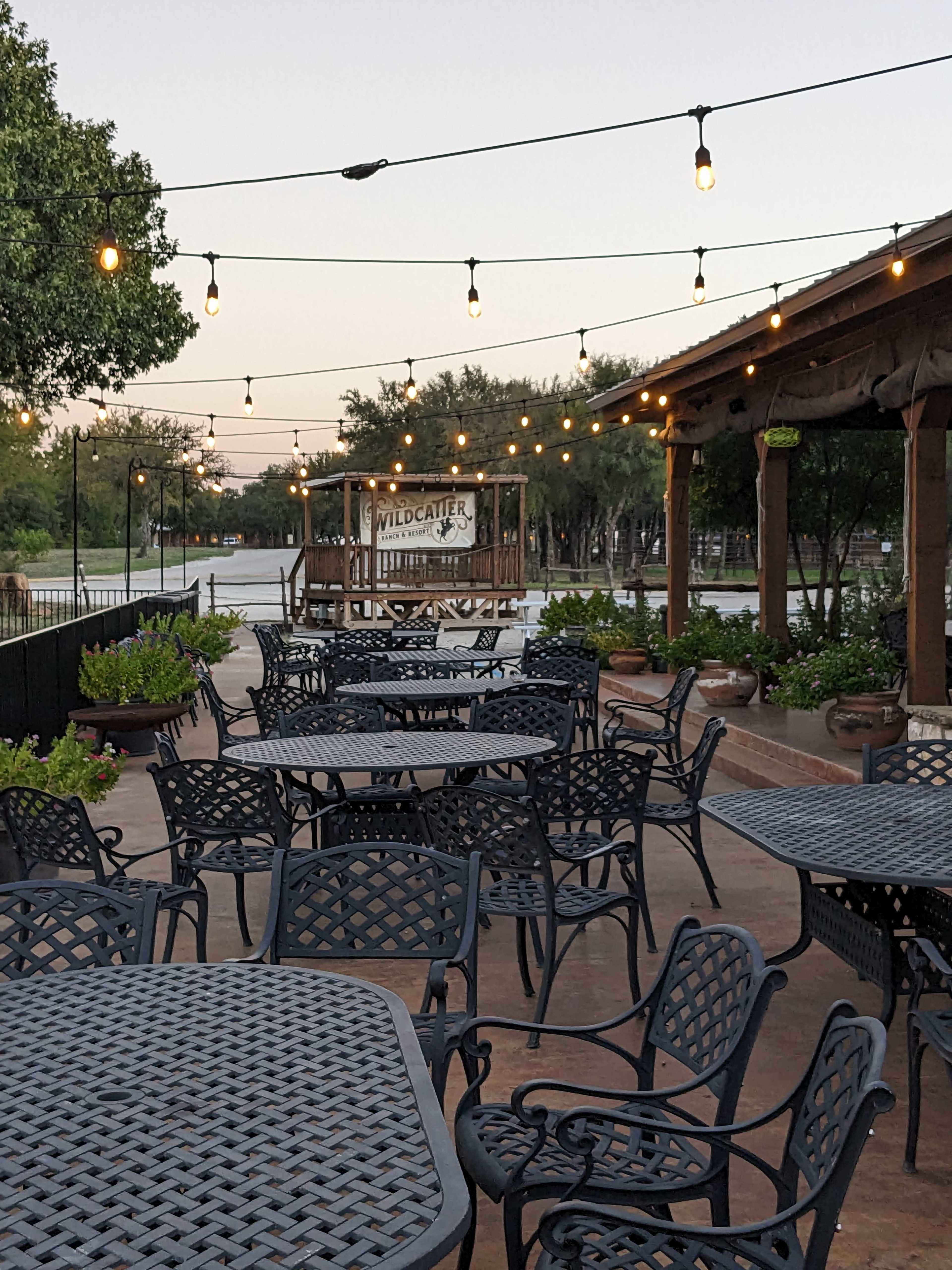 The image shows a patio area of a restaurant with black metal tables and chairs, string lights overhead, and a view of a sign that reads "WILDCATTERS" in the background.