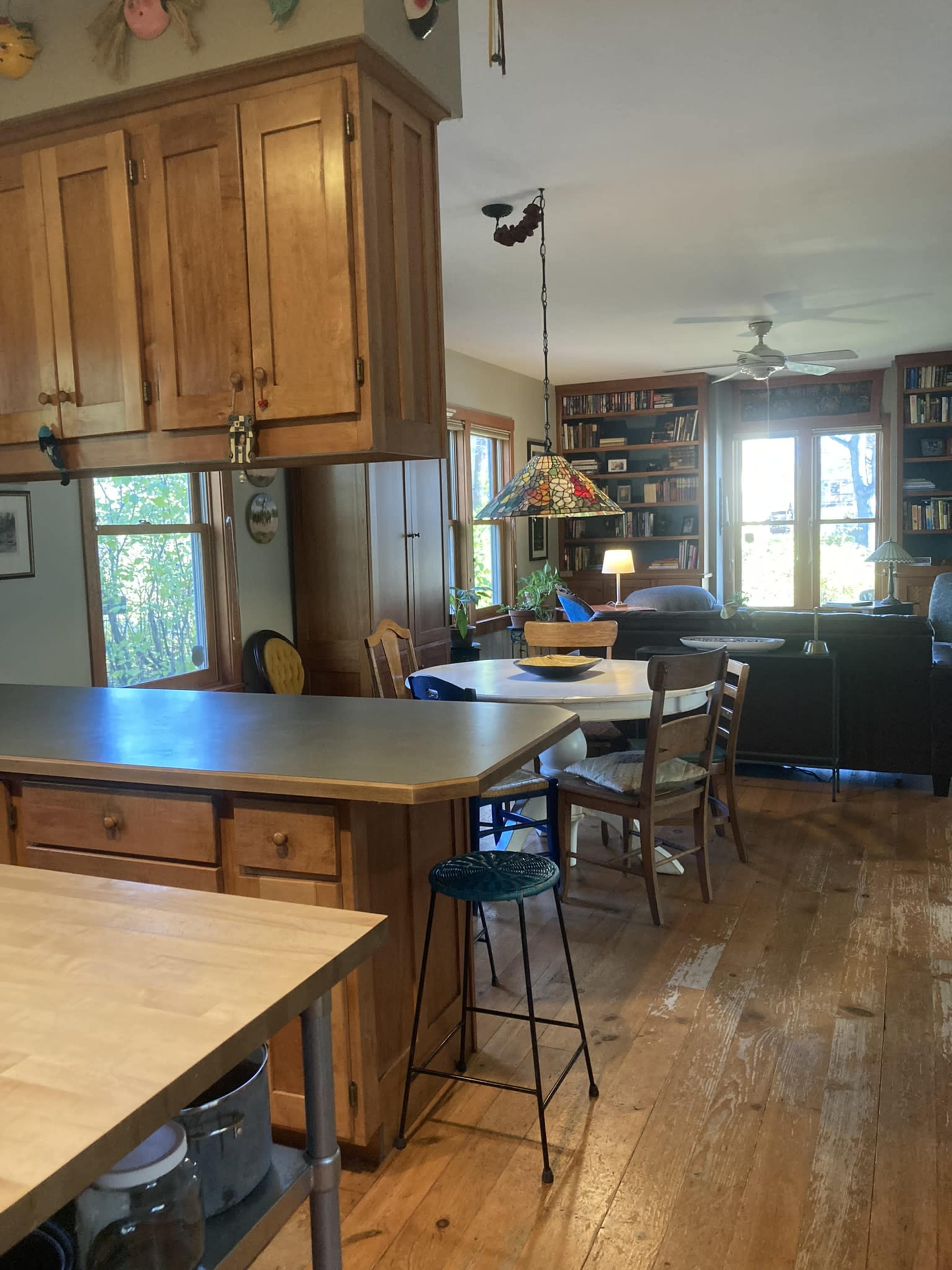 A wooden kitchen with a central island, a dining table surrounded by chairs, and a living area visible through large windows.