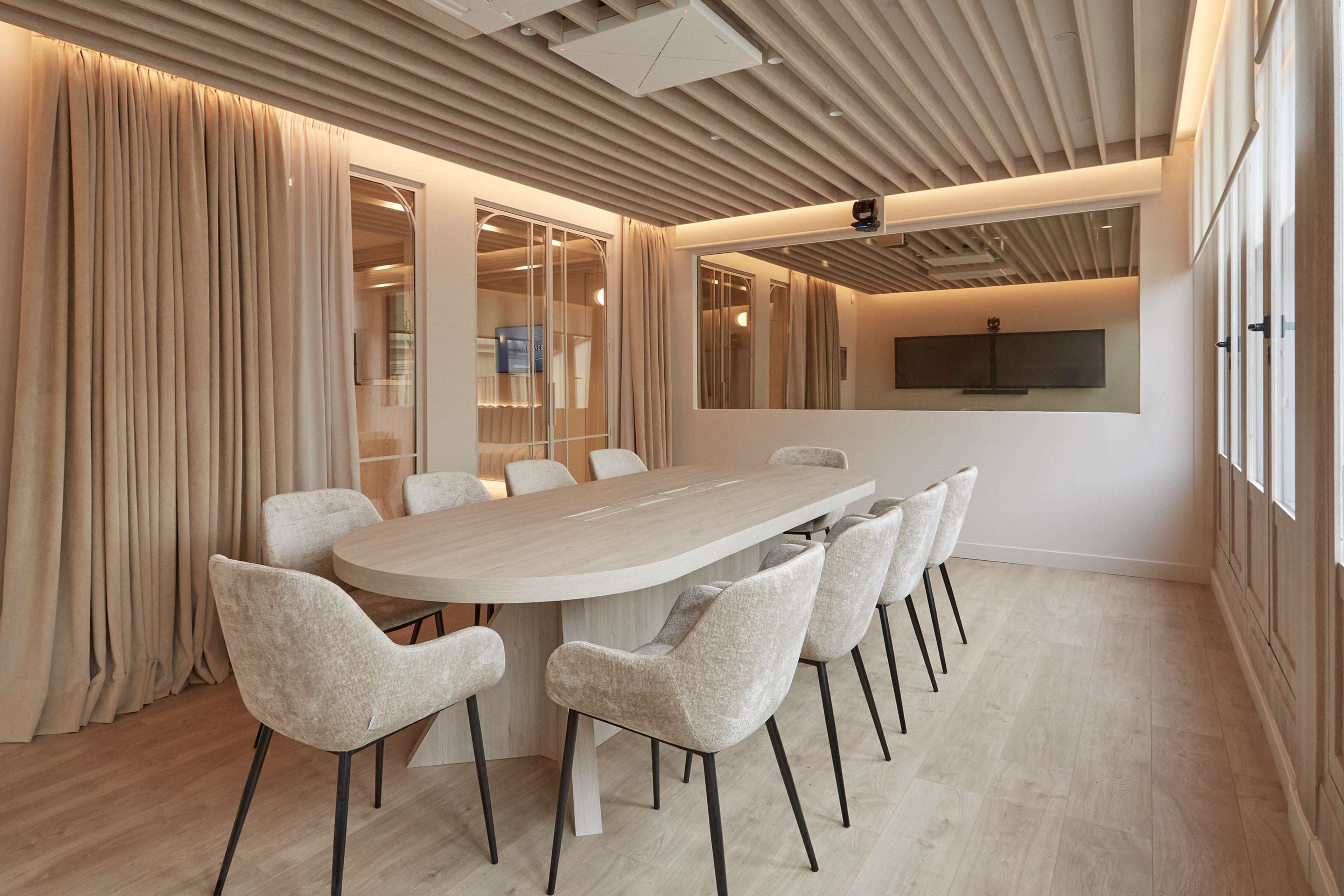 A modern conference room features a large oval table surrounded by upholstered chairs, with soft lighting and curtain-covered windows.