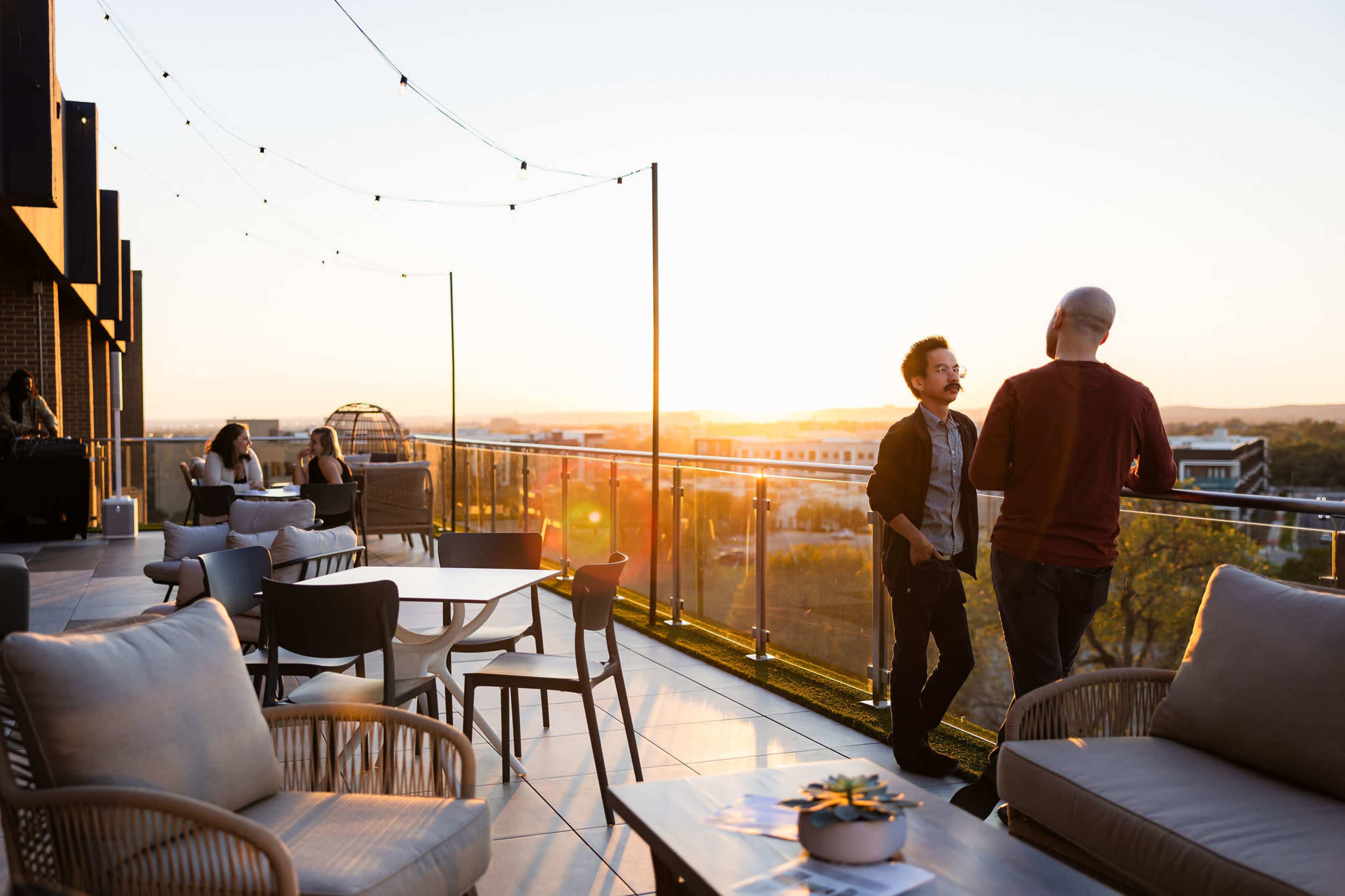 The image shows a rooftop terrace at sunset with people engaged in conversation and seating arranged around tables.