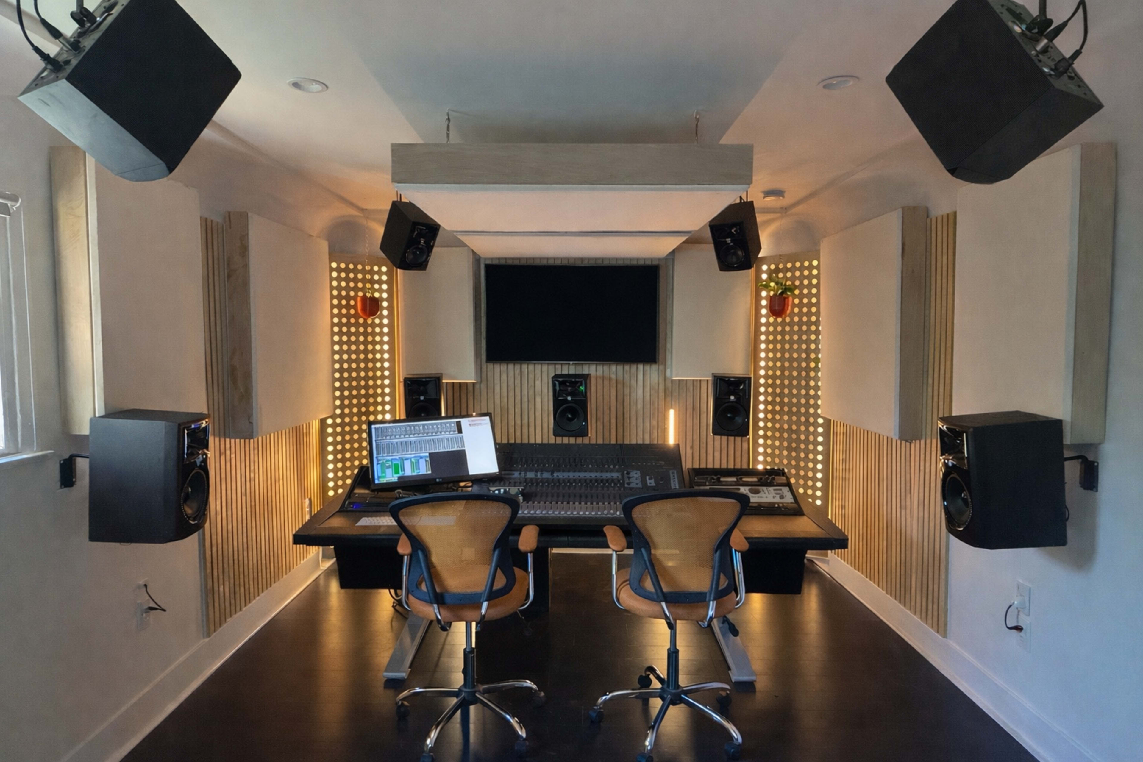 The image shows a modern recording studio featuring a central mixing desk, two office chairs, and audio equipment, with soundproofing panels and an ambient light setup on the walls.
