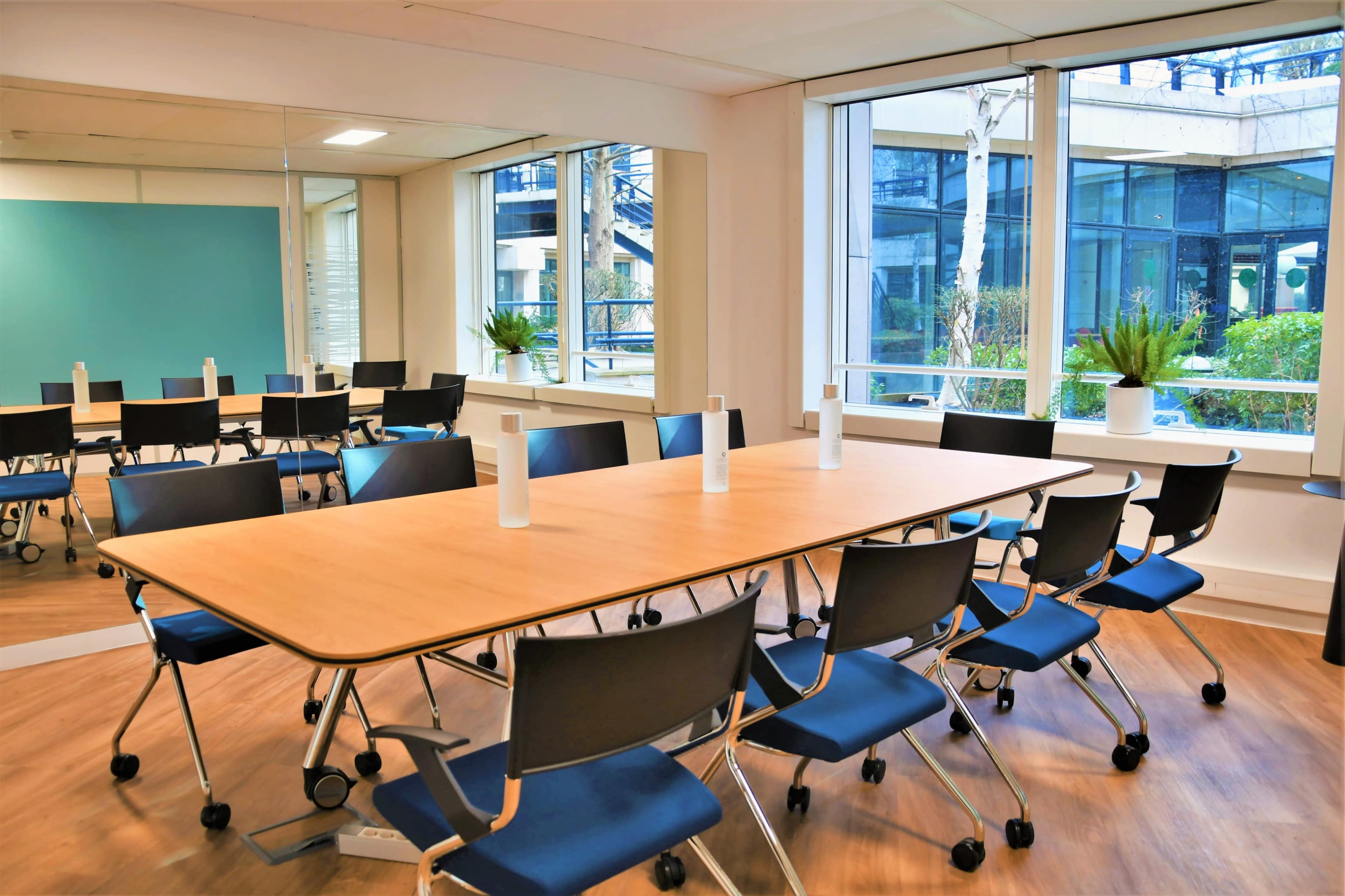 A modern conference room with a long wooden table surrounded by black rolling chairs, large windows, and greenery visible outside.