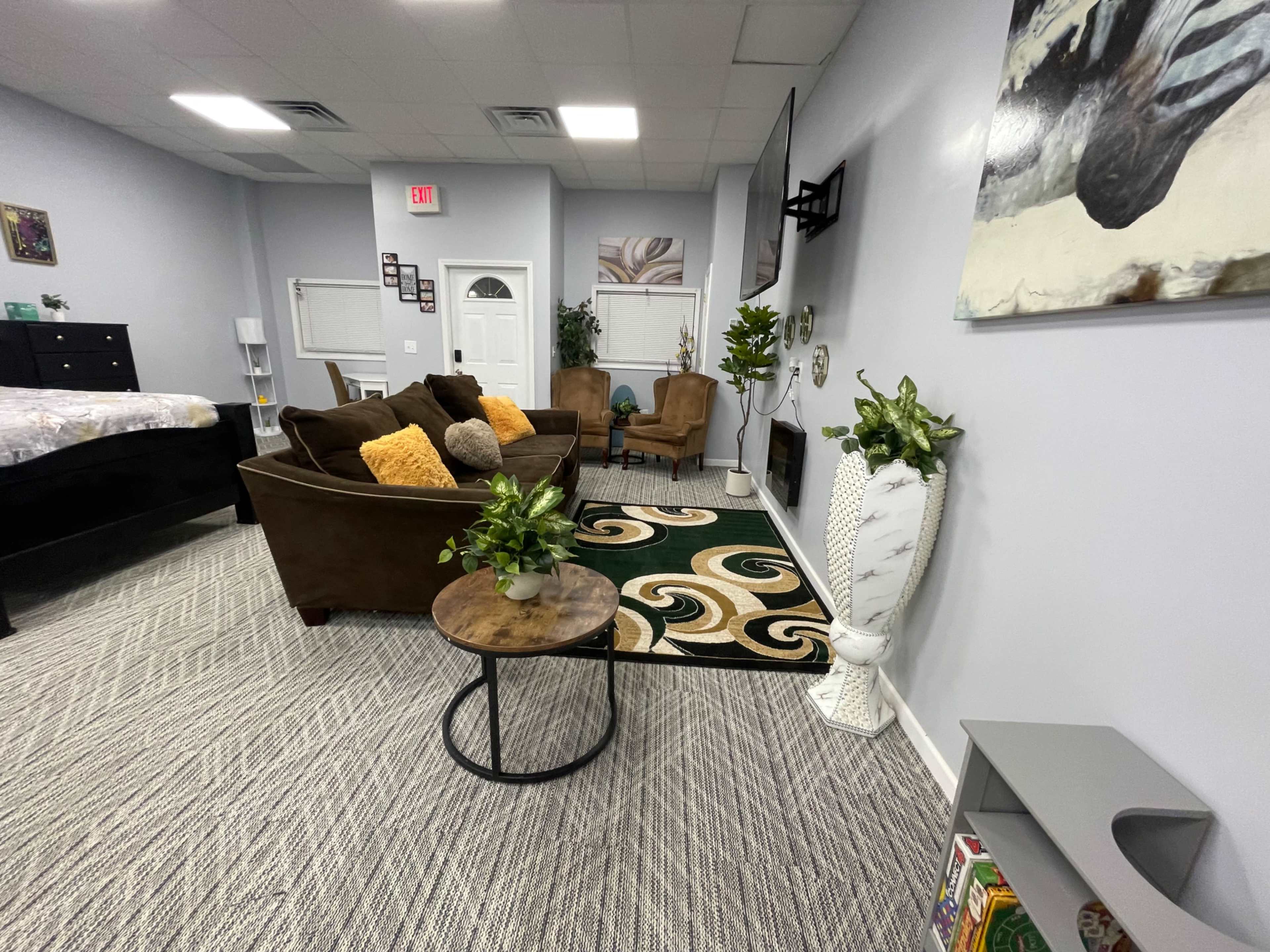 The image shows a cozy room with a brown sectional sofa, a round coffee table, and decorative plants, alongside a bed and a television on the wall.