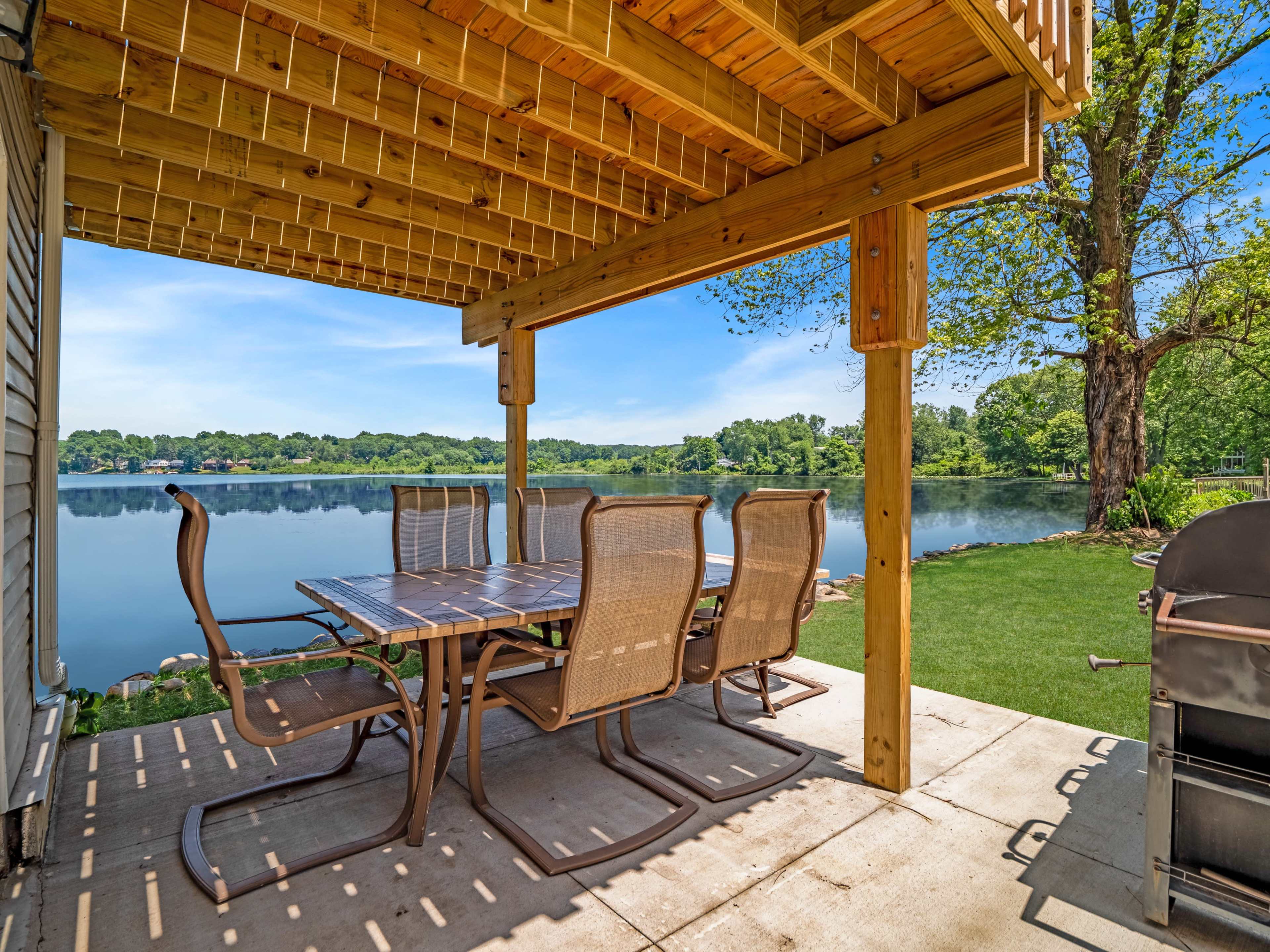 A patio area with a large table and chairs overlooks a calm lake, framed by a wooden pergola and tree.