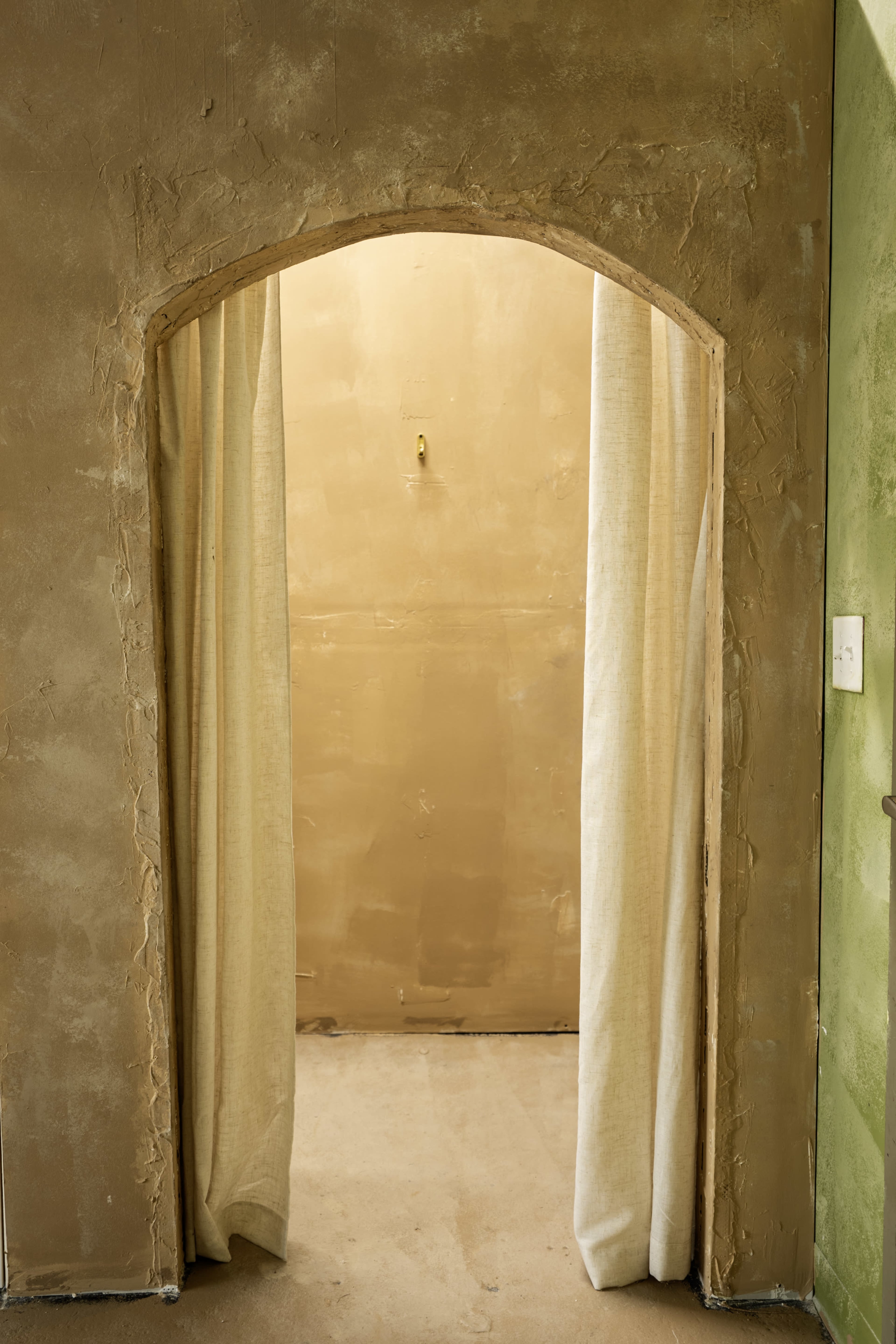 The image shows an archway with beige curtains leading into a simple, unadorned room with light-colored walls and a bare floor.