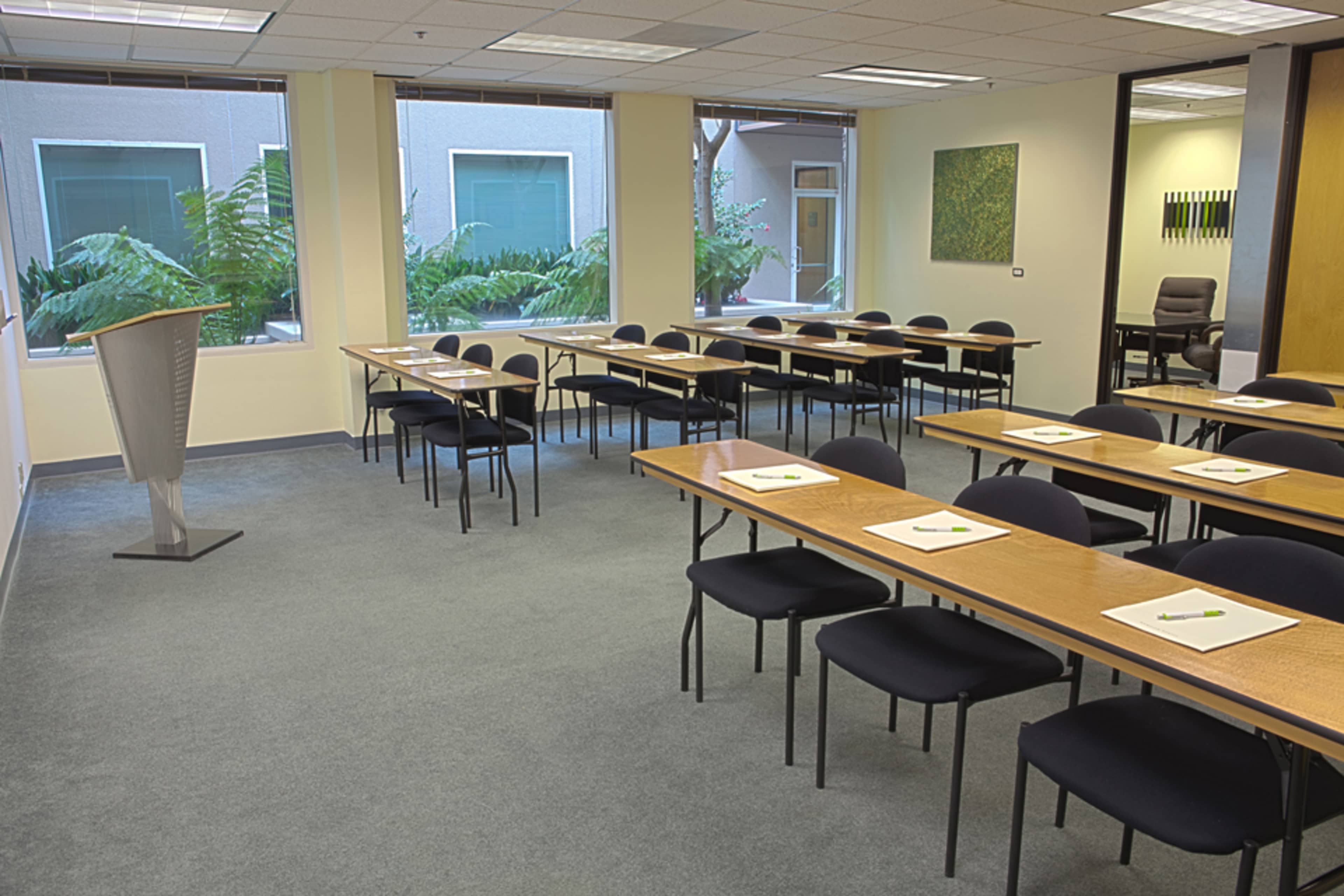 A conference room is set up with multiple tables and chairs, facing a podium, and features large windows overlooking a green courtyard.