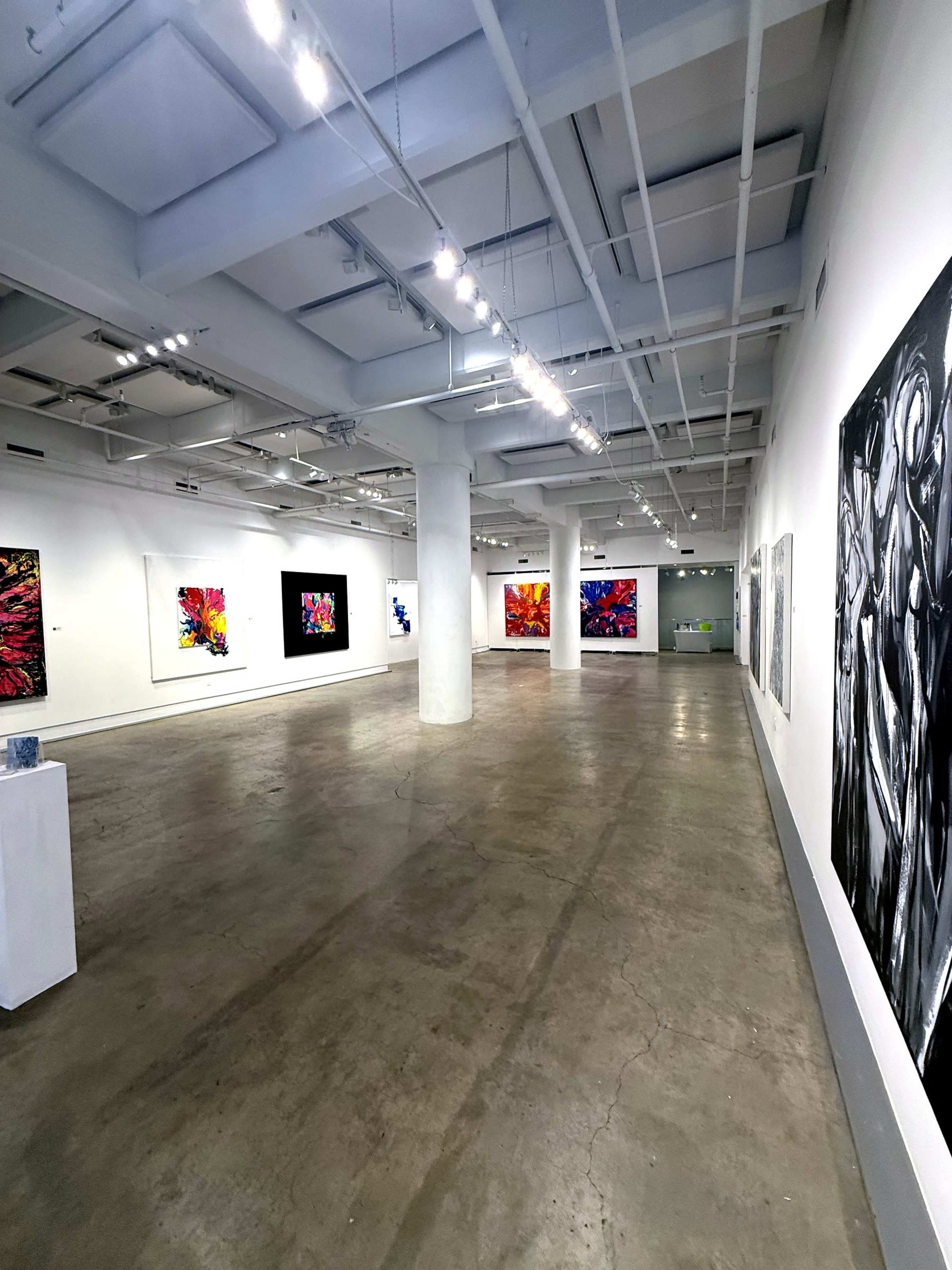 The image shows a spacious art gallery featuring vibrant abstract paintings on the white walls and a polished concrete floor.