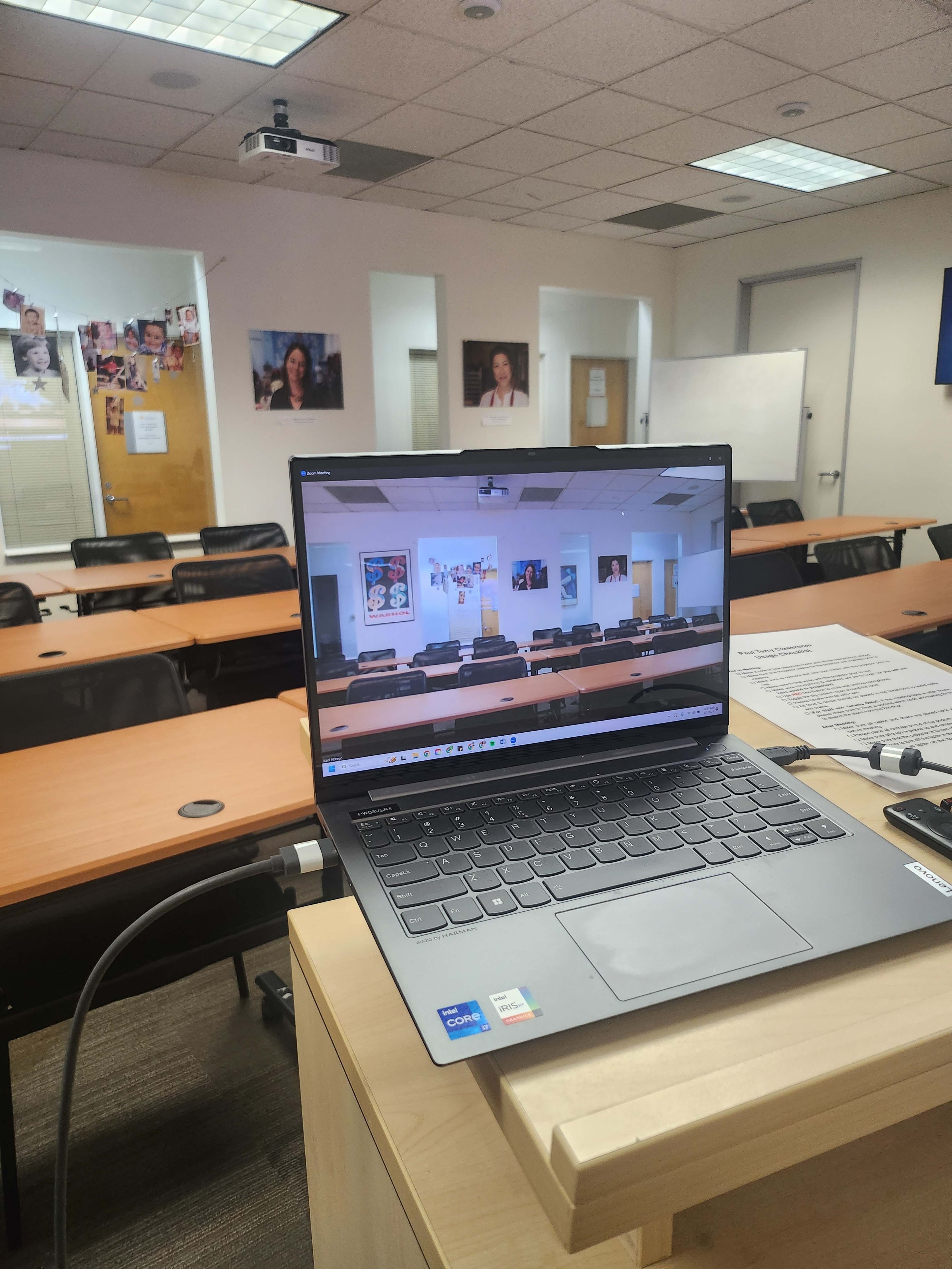 A laptop displaying a classroom scene is placed on a table in an empty conference room with chairs arranged around it.