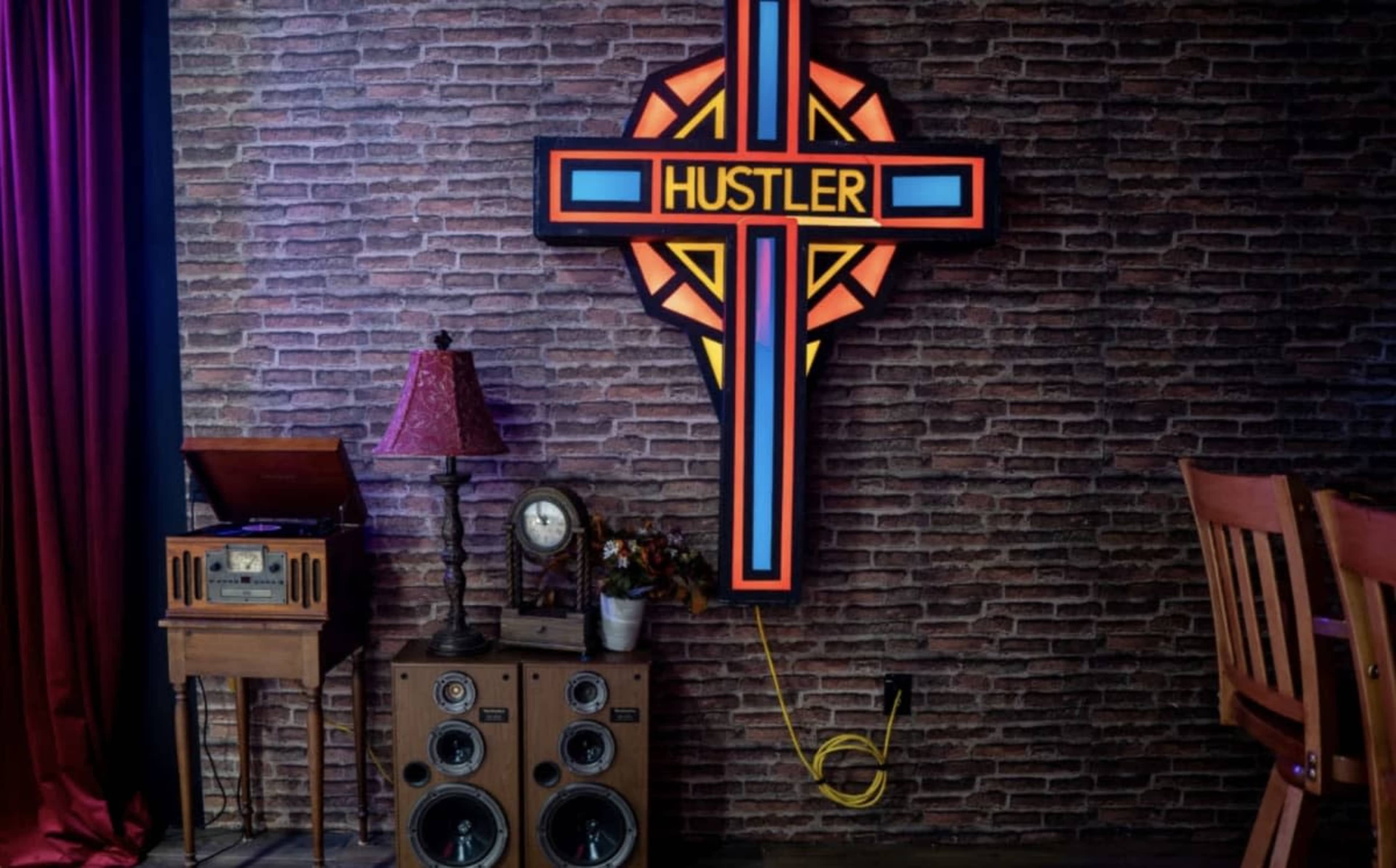 The image features a neon sign reading "HUSTLER" mounted on a brick wall, accompanied by vintage furniture including a record player, a lamp, and a stereo system.