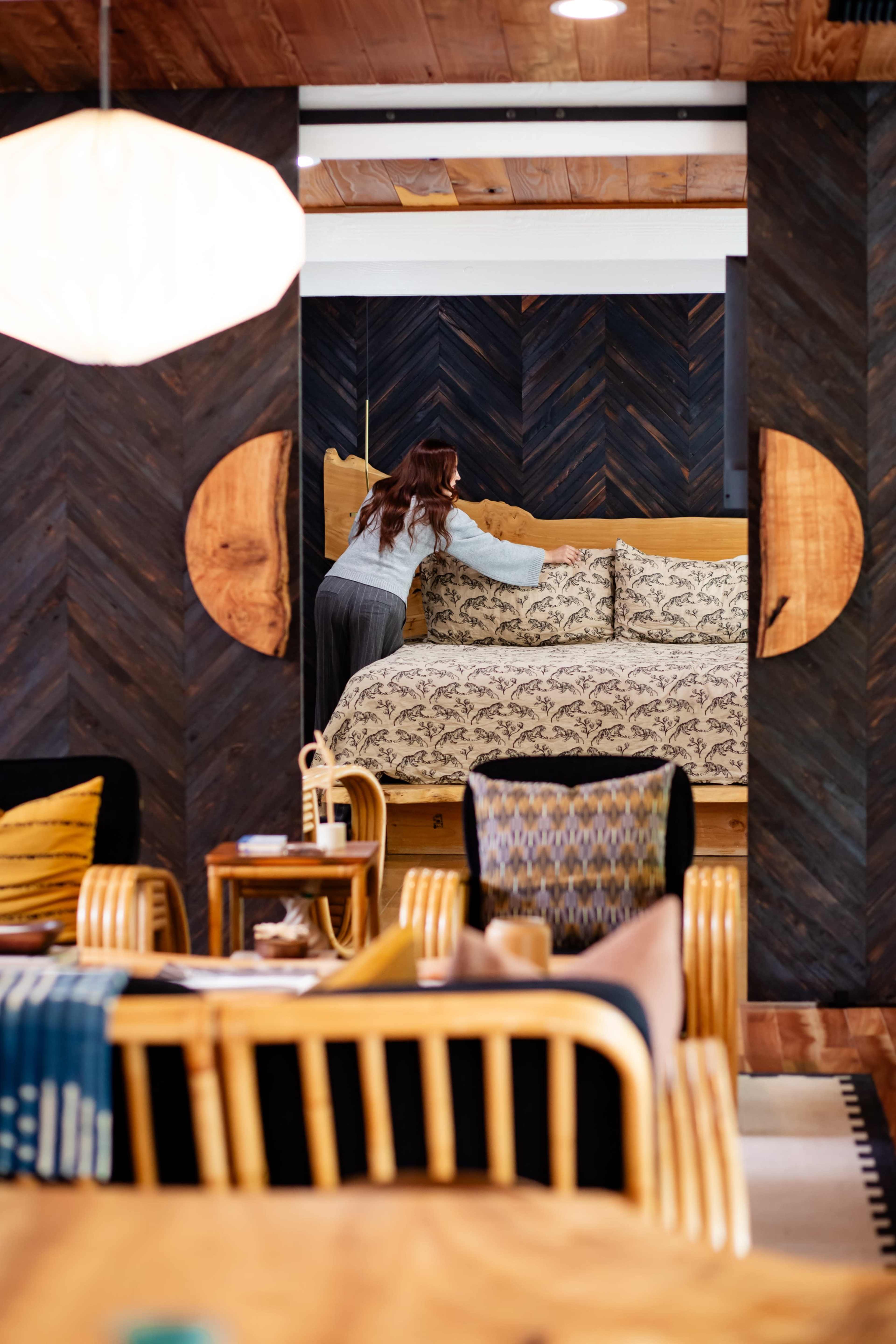 A woman arranges pillows on a bed in a stylishly decorated room featuring dark wood and modern furnishings.