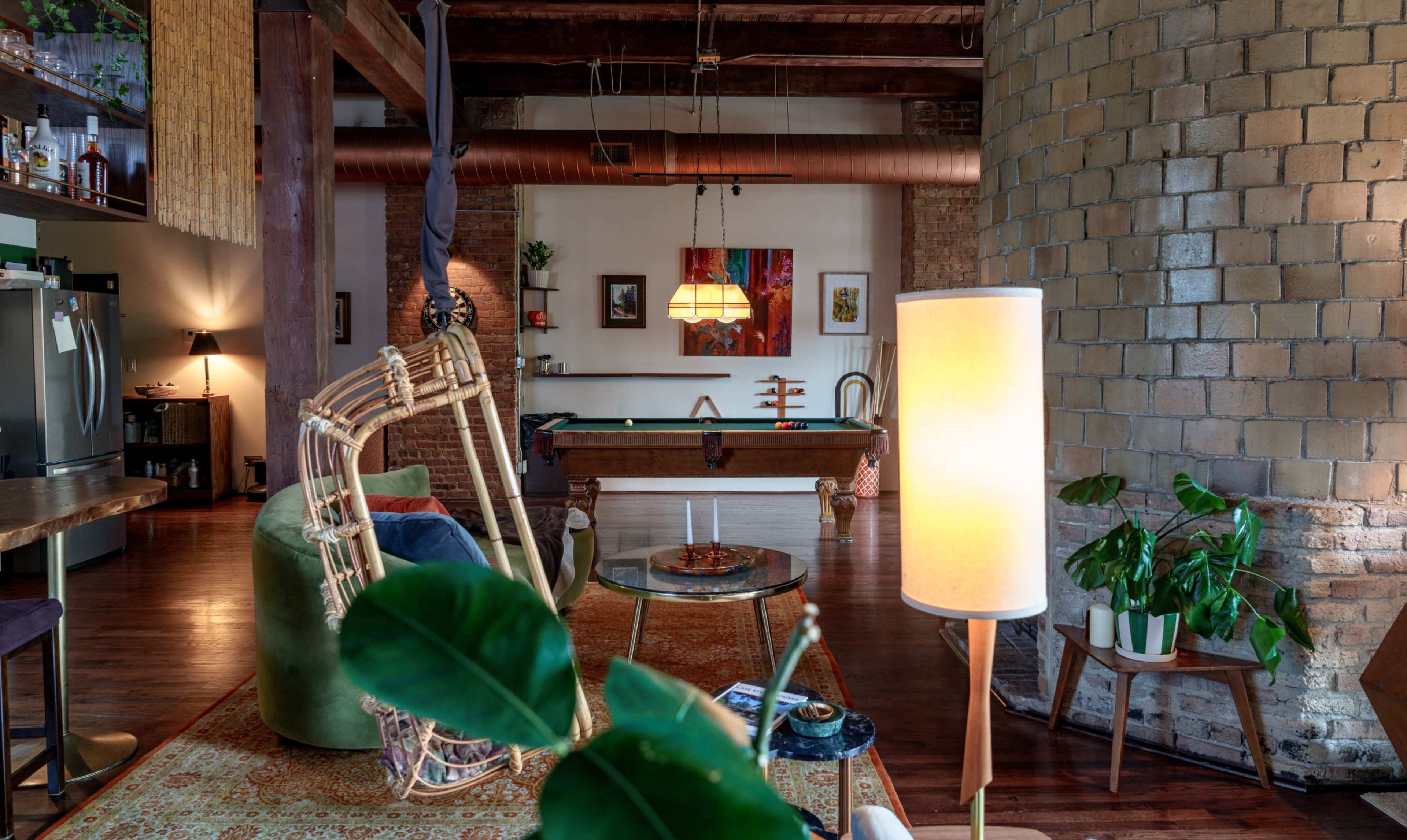 The image depicts an open-concept living space with exposed brick walls, a pool table in the background, and various seating arrangements complemented by plants and decorative elements.