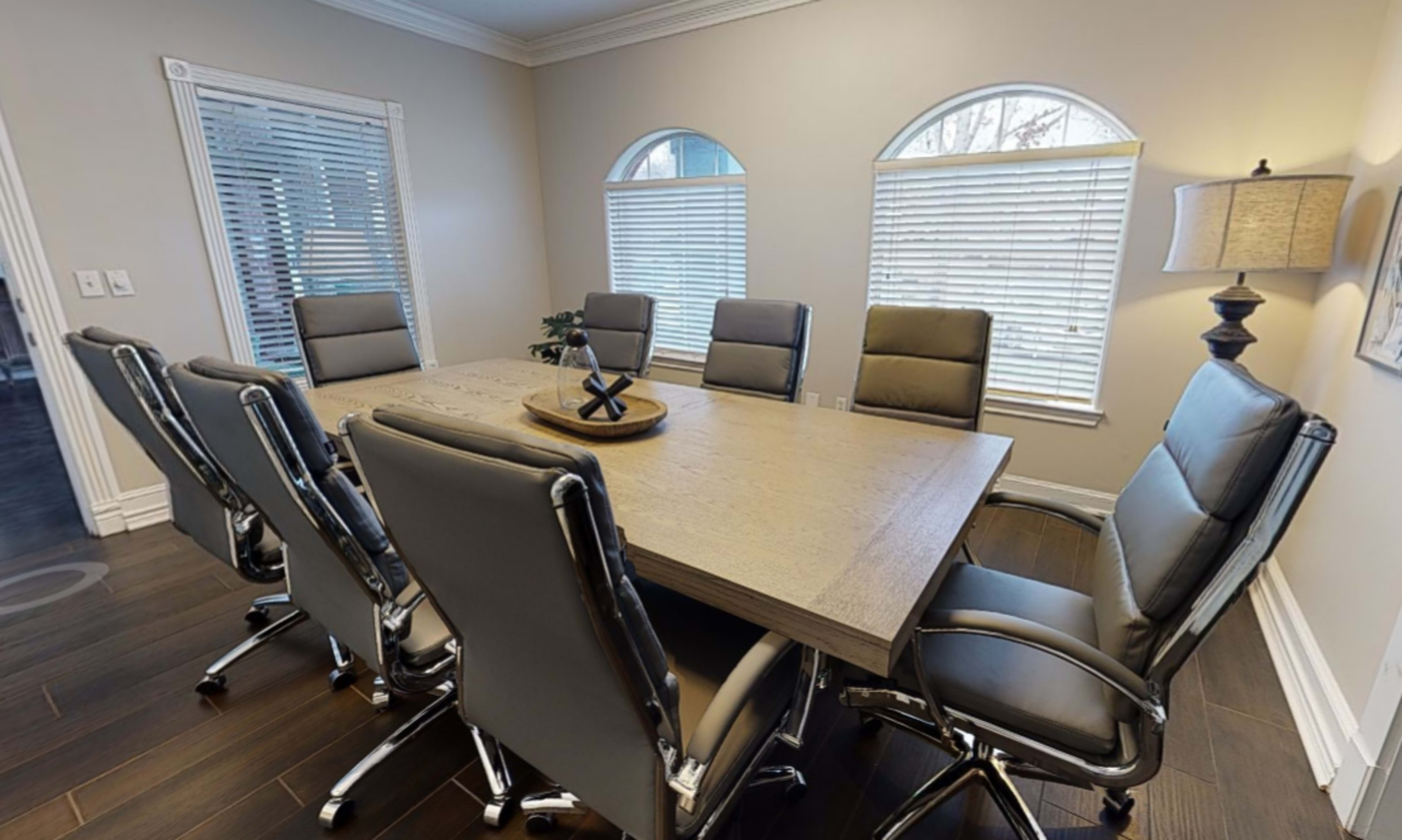 A large conference table surrounded by eight ergonomic chairs in a well-lit meeting room with arched windows.