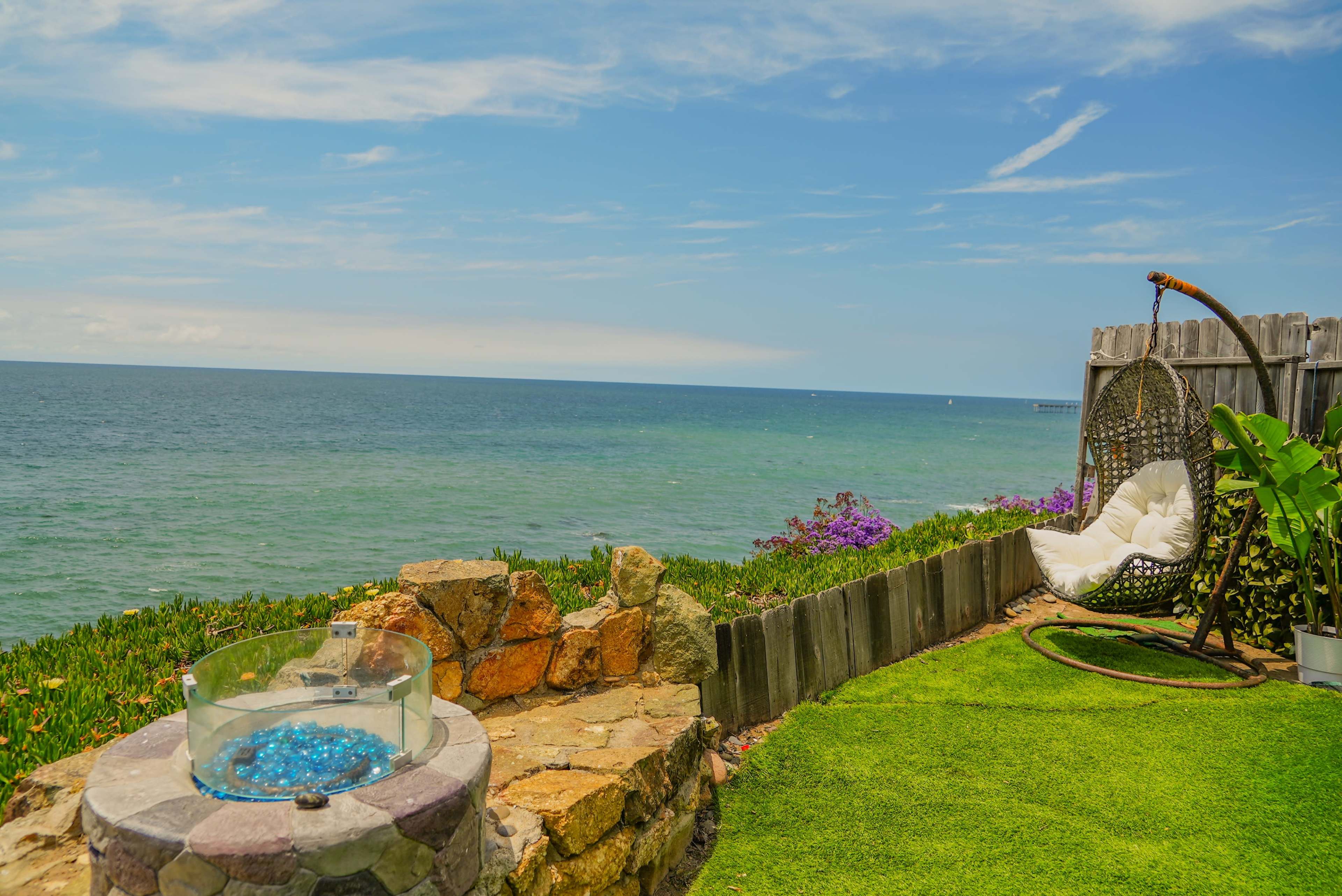 A coastal patio features a stone seating area, a chair suspended from a frame, and a view of the ocean beyond.