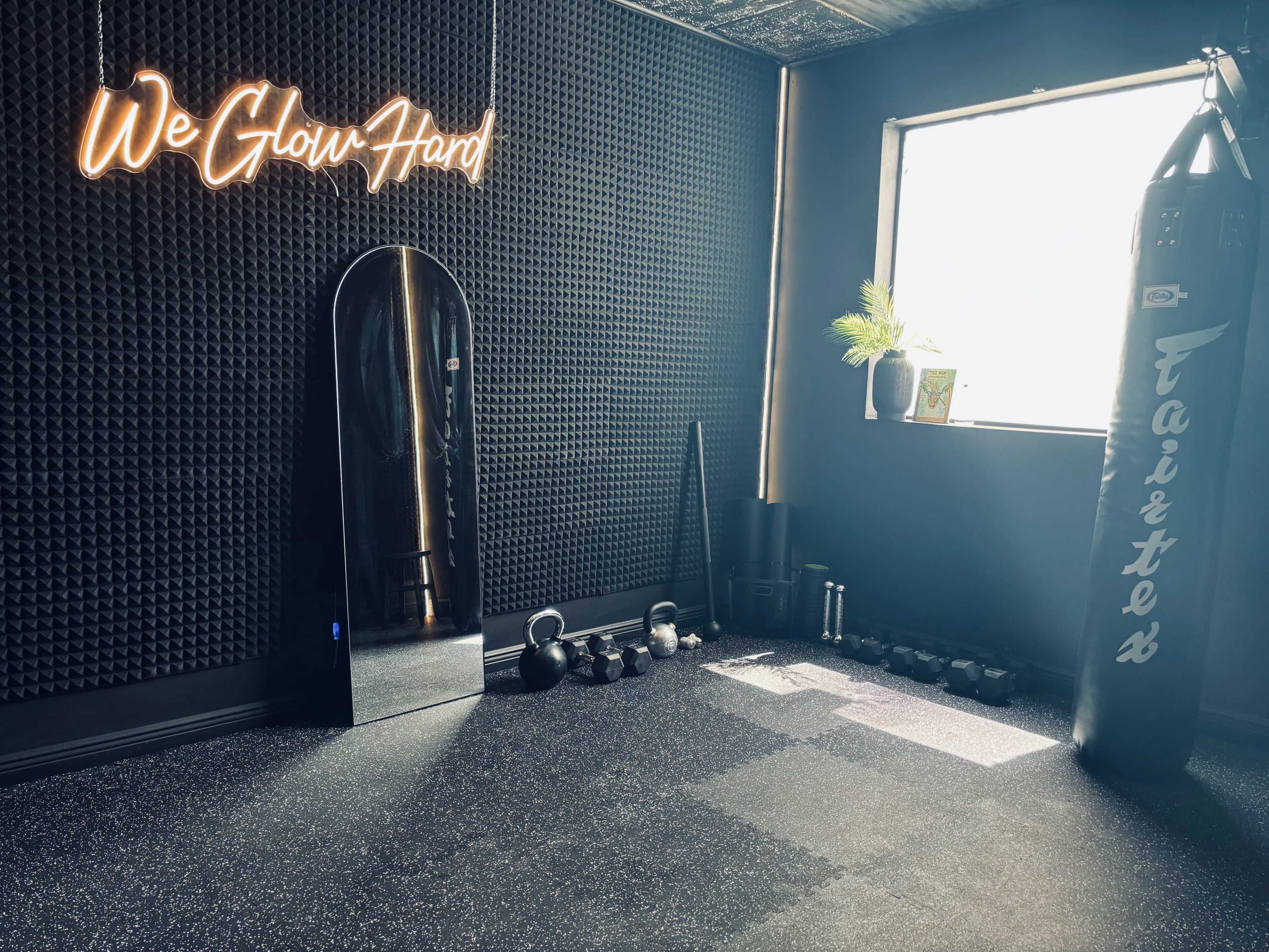 A fitness room with a neon sign reading "We Glow Hard," a large mirror, exercise equipment, a punching bag, and a window allowing natural light.