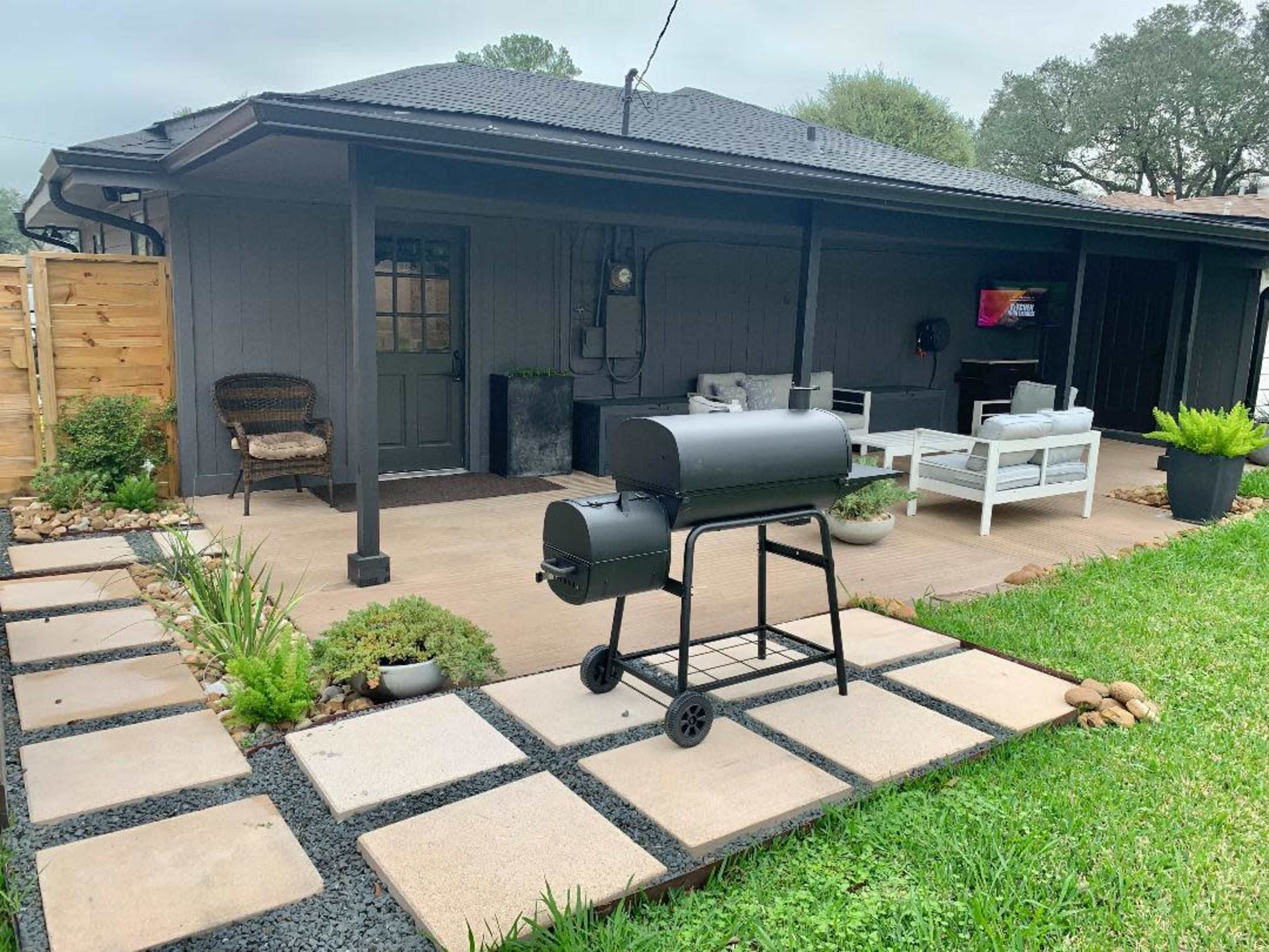 A backyard patio with a charcoal grill on a stone walkway, surrounded by greenery and outdoor furniture.