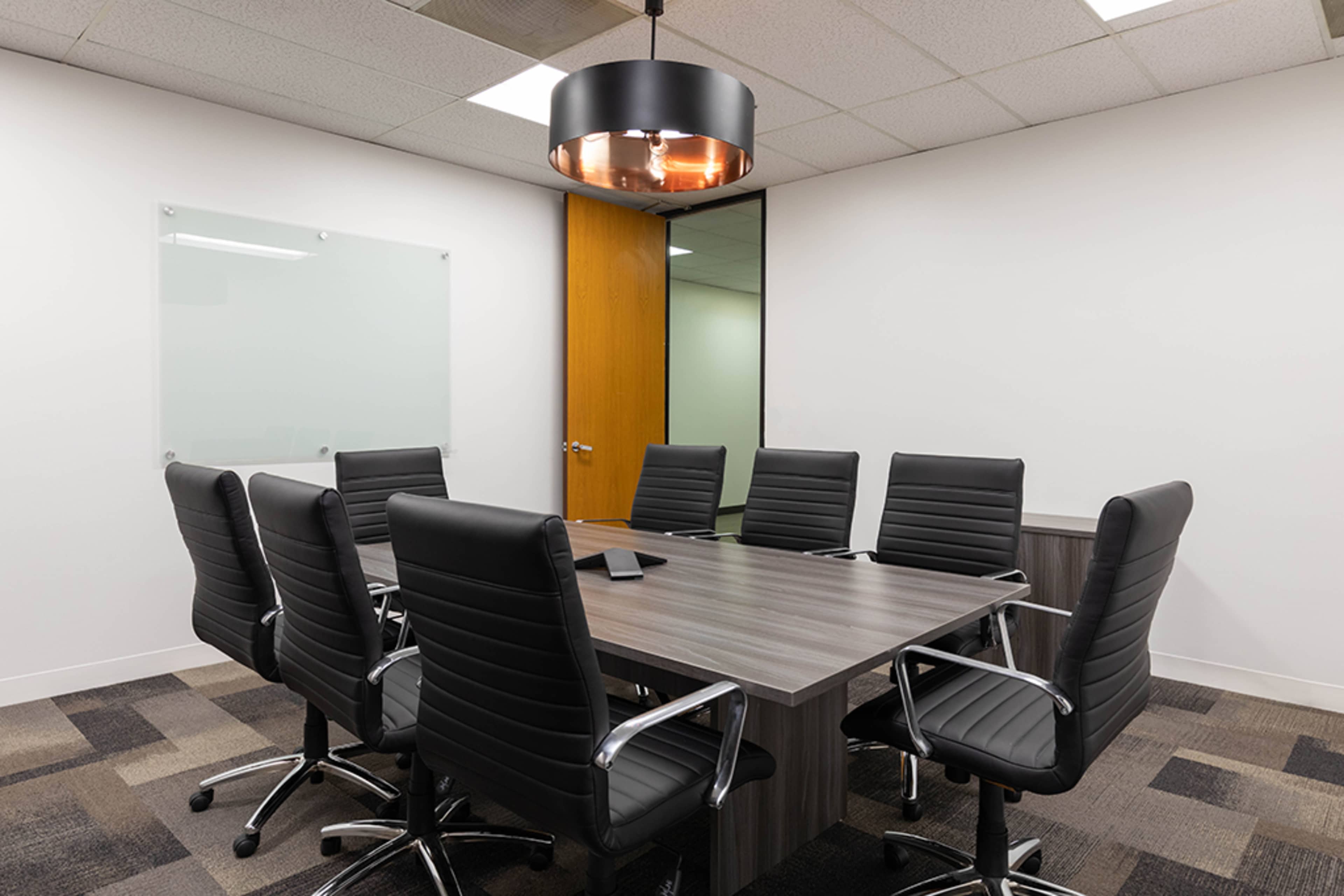 A conference room features a large wooden table surrounded by eight black swivel chairs under a circular pendant light.