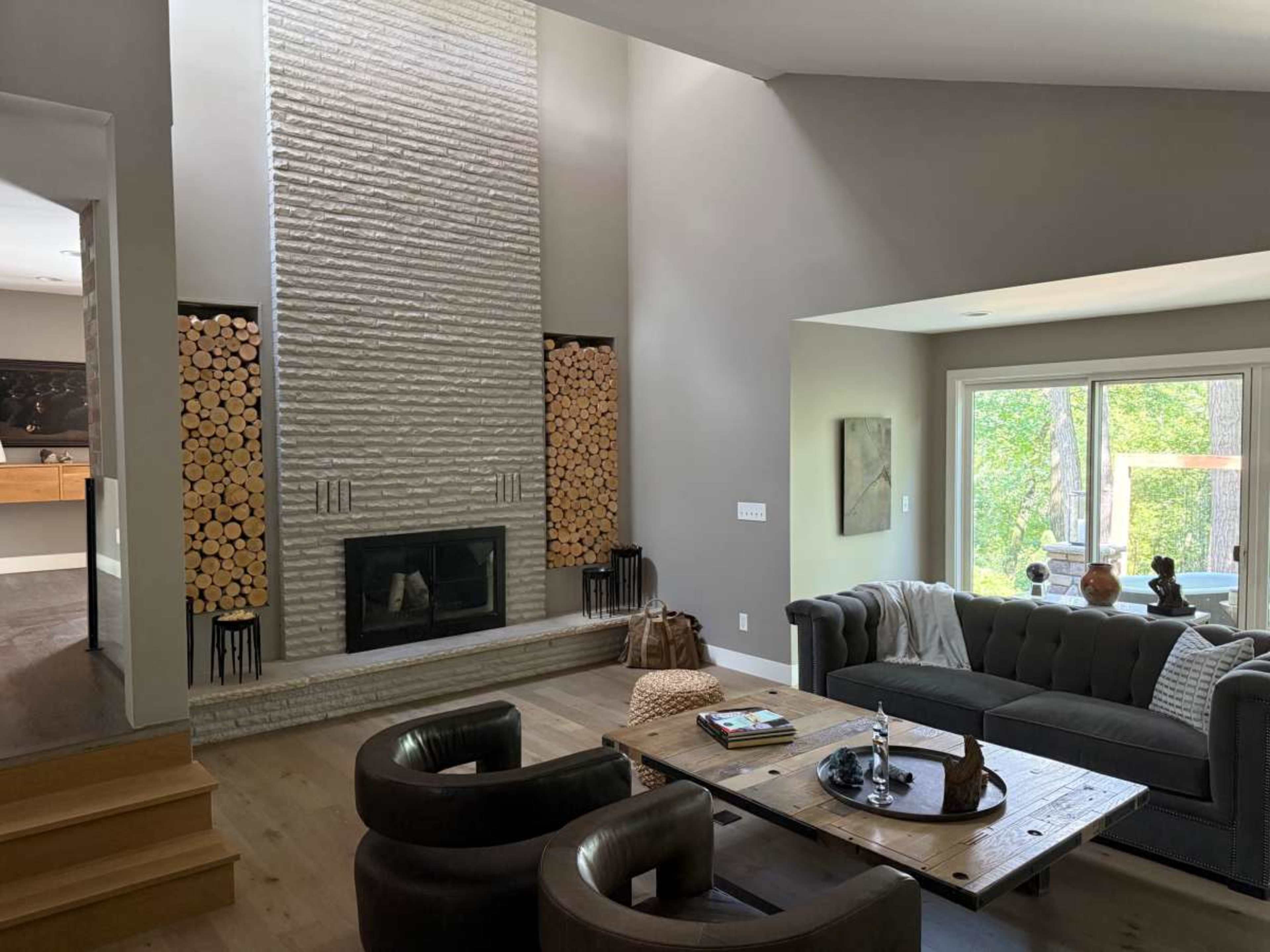 The image shows a modern living room featuring a fireplace with stacked firewood, a large window displaying greenery outside, and comfortable seating arranged around a wooden coffee table.