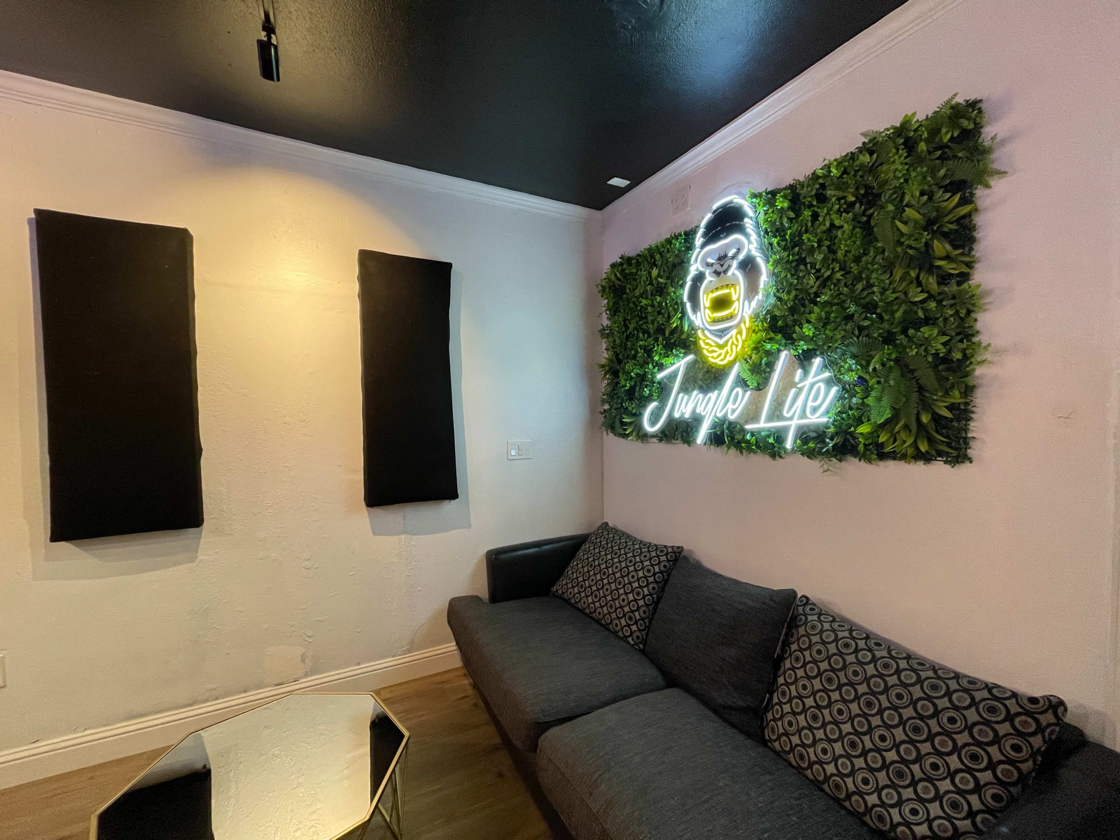 The image shows a cozy lounge area featuring a black sofa, a geometric coffee table, and a green wall decoration with a neon sign reading "Jungle Life."