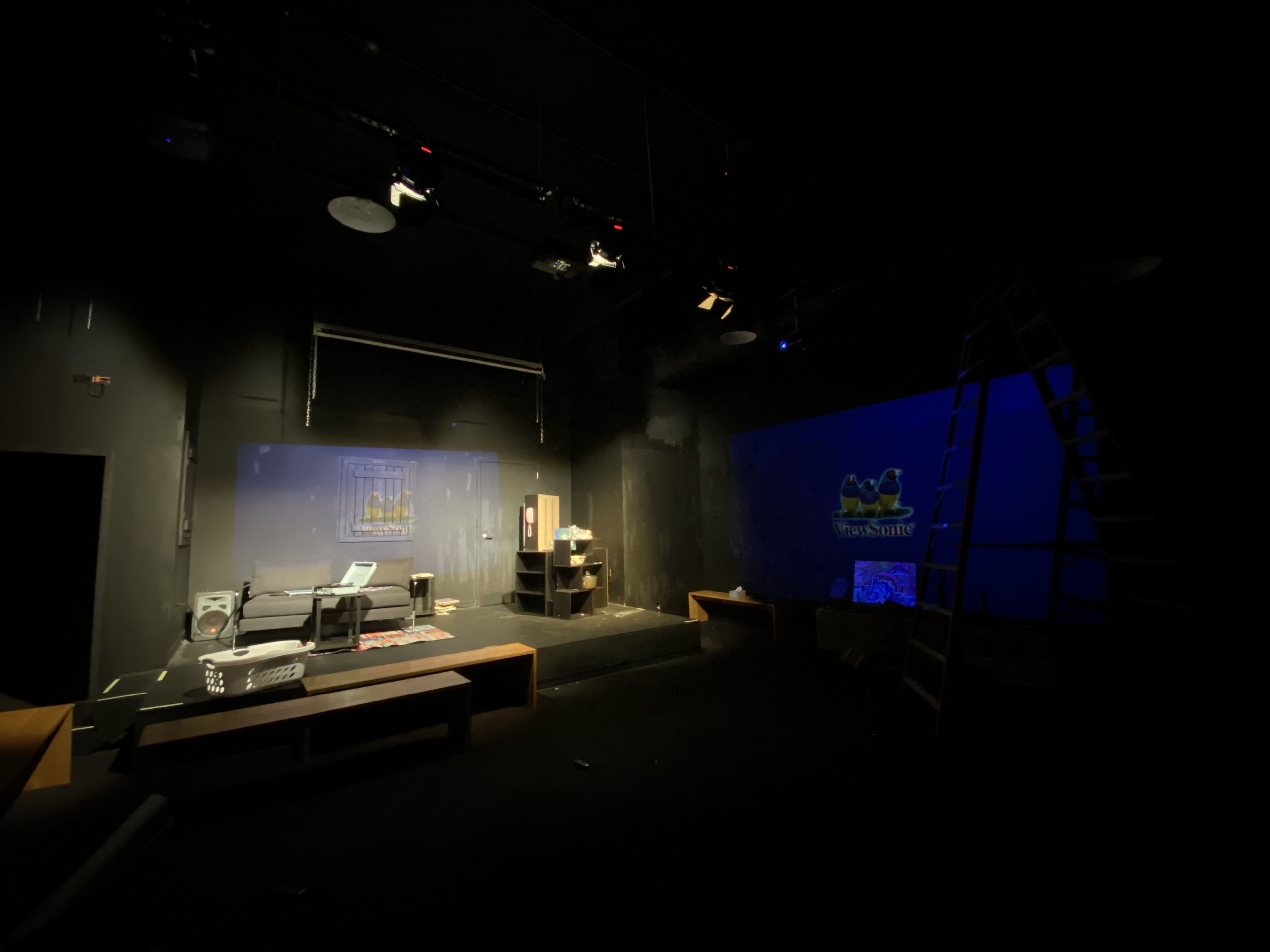 A dimly lit theater stage with a set featuring a couch, shelves, and a ladder, along with a blue backdrop and stage lighting equipment.