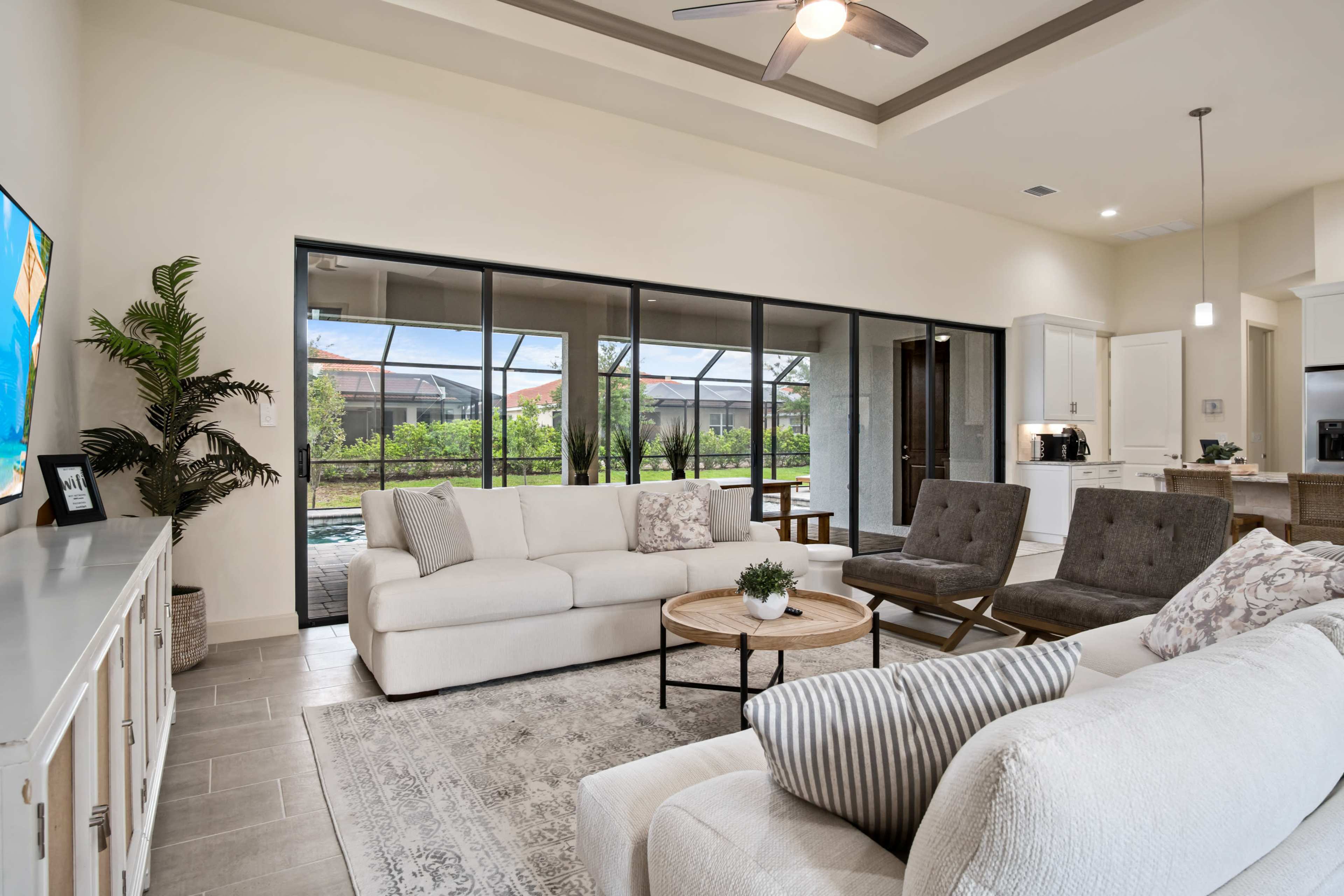 A modern living room featuring a white sofa, gray armchairs, a round coffee table, and large windows overlooking a patio and garden area.