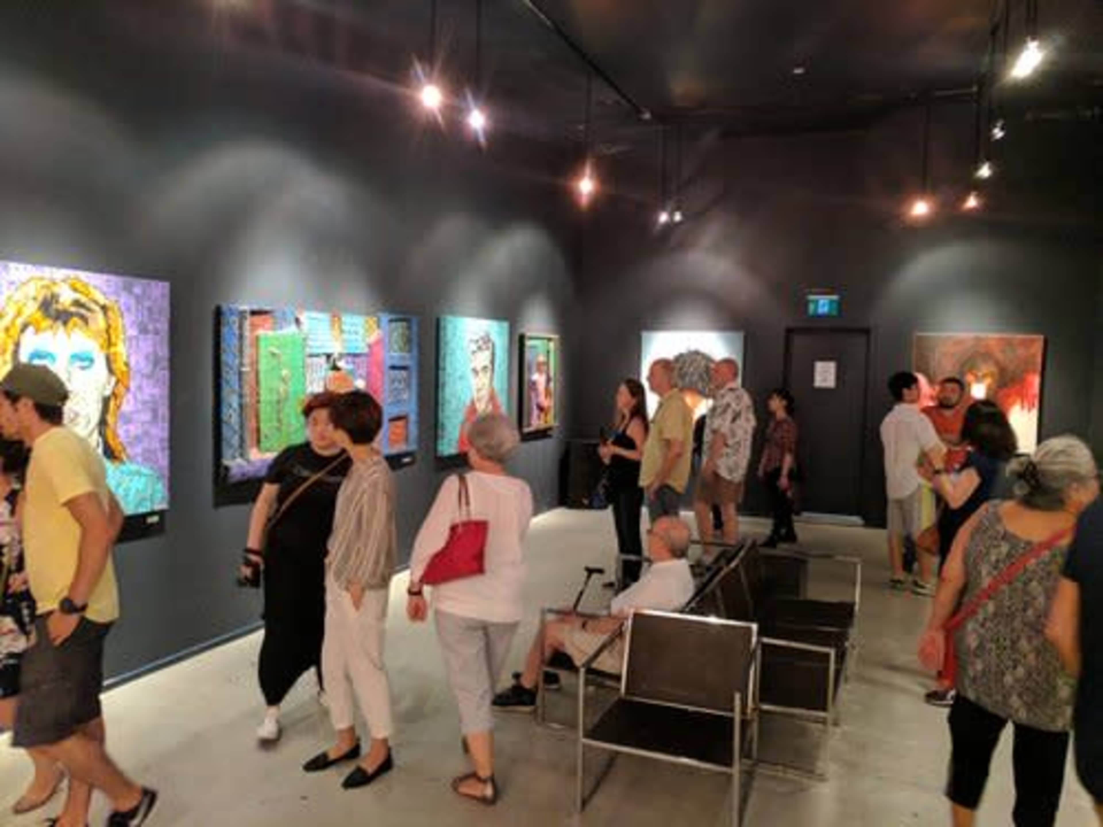 A crowded art gallery features various colorful paintings on the walls while visitors chat and observe the exhibits.