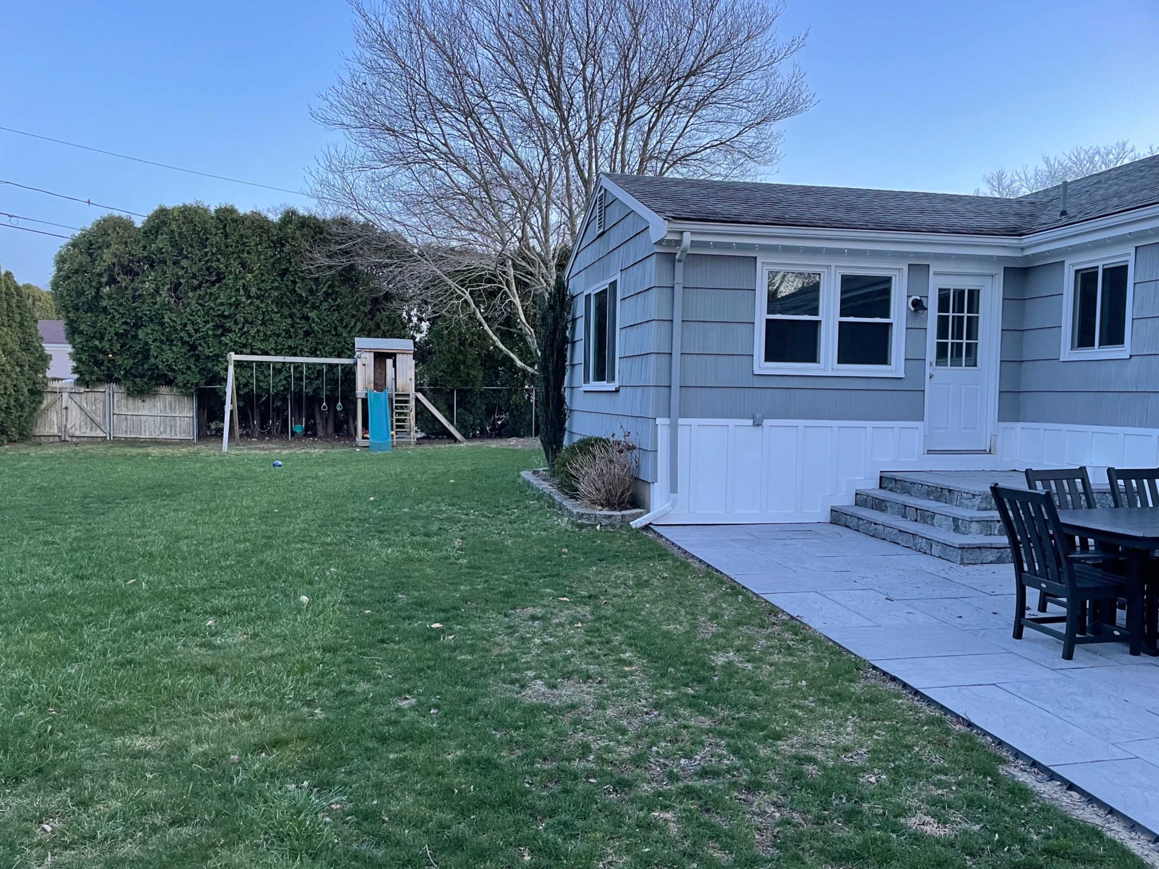 A backyard features a patio area with a table and chairs adjacent to a playset and a grassy lawn.