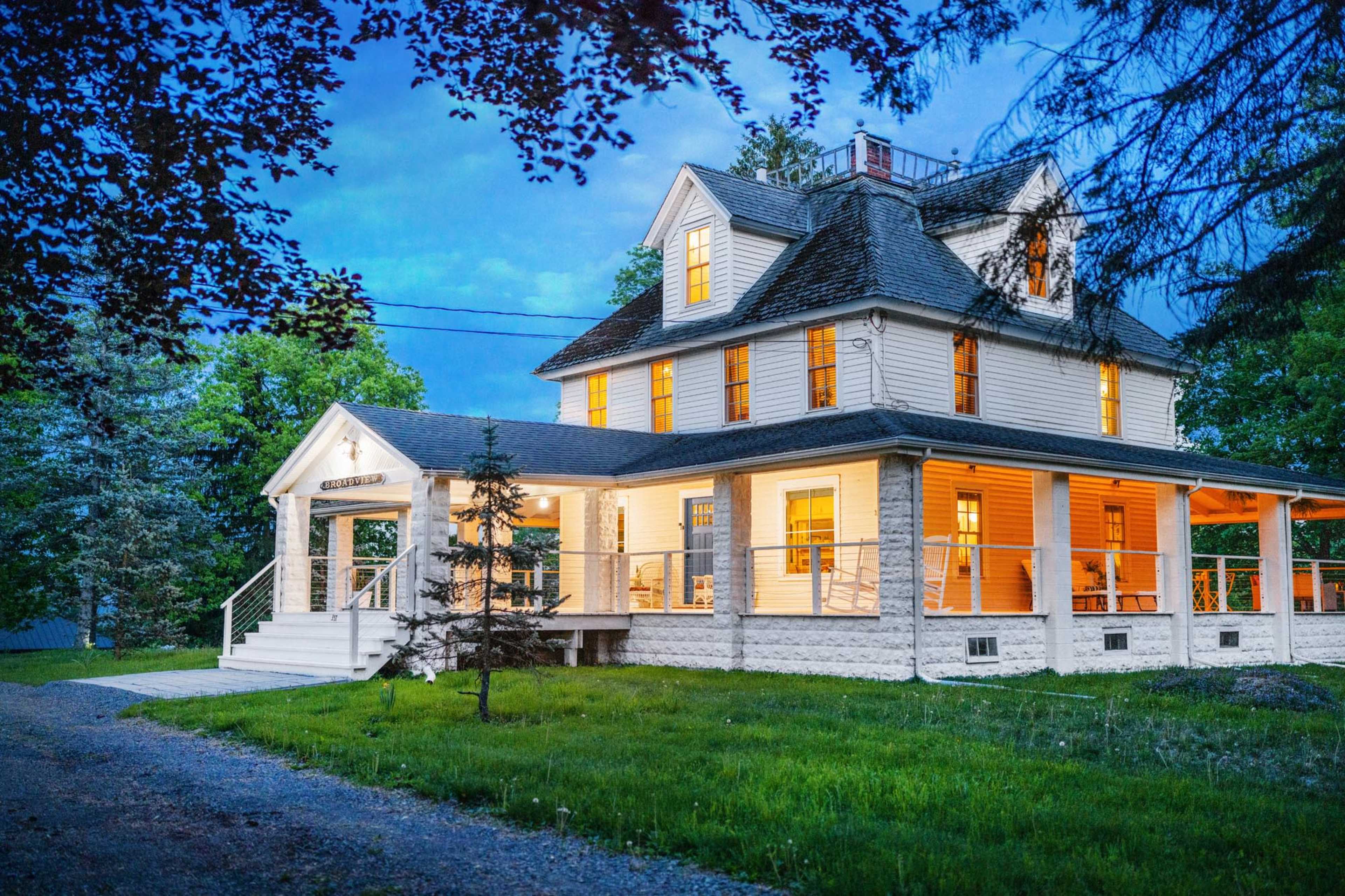 A two-story white house with a porch, surrounded by green grass and trees, illuminated in the evening light.