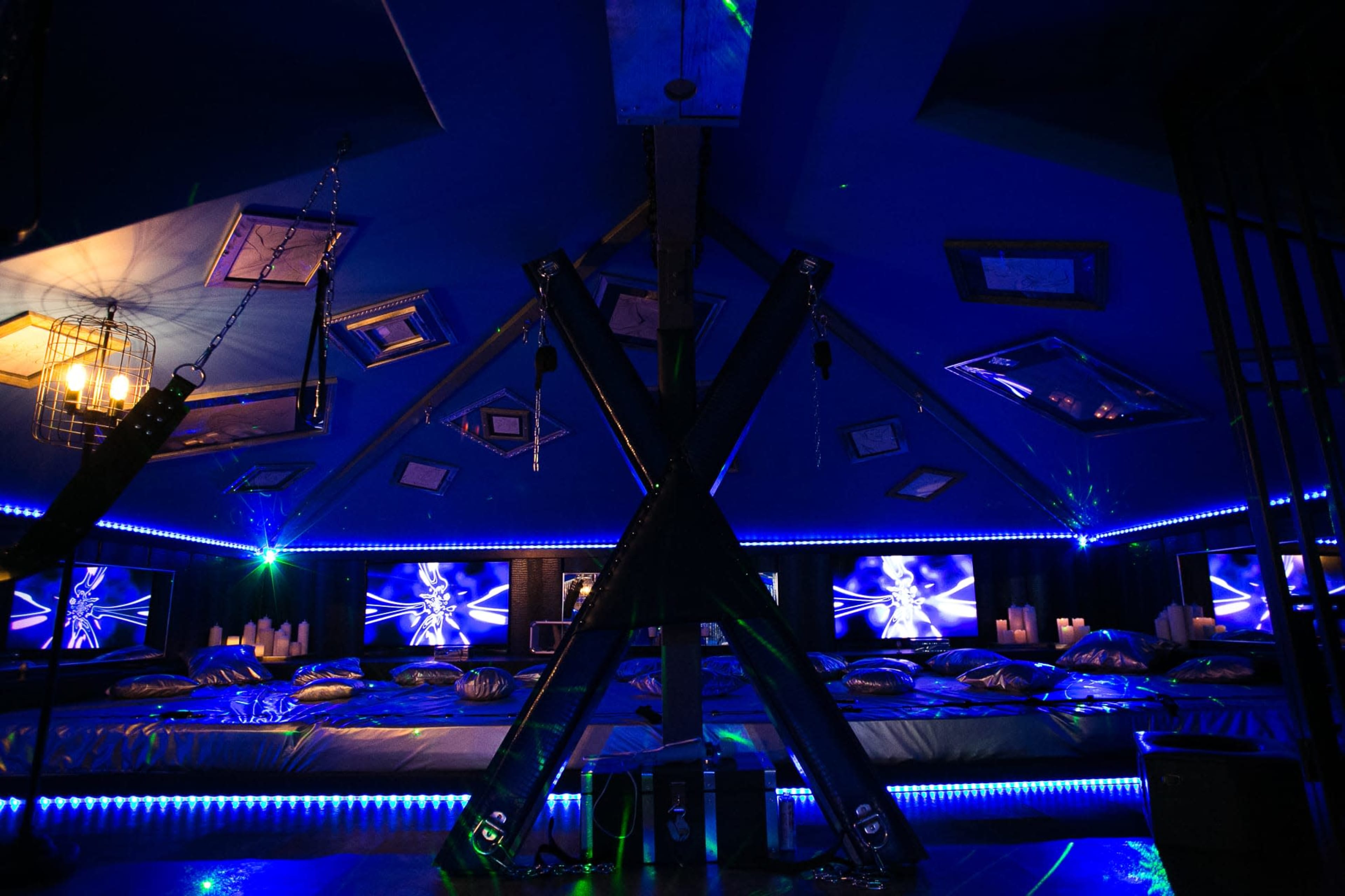 A dimly lit room with a triangular ceiling, featuring blue LED lights, multiple framed pictures on the walls, and a seating area with cushions and screens.