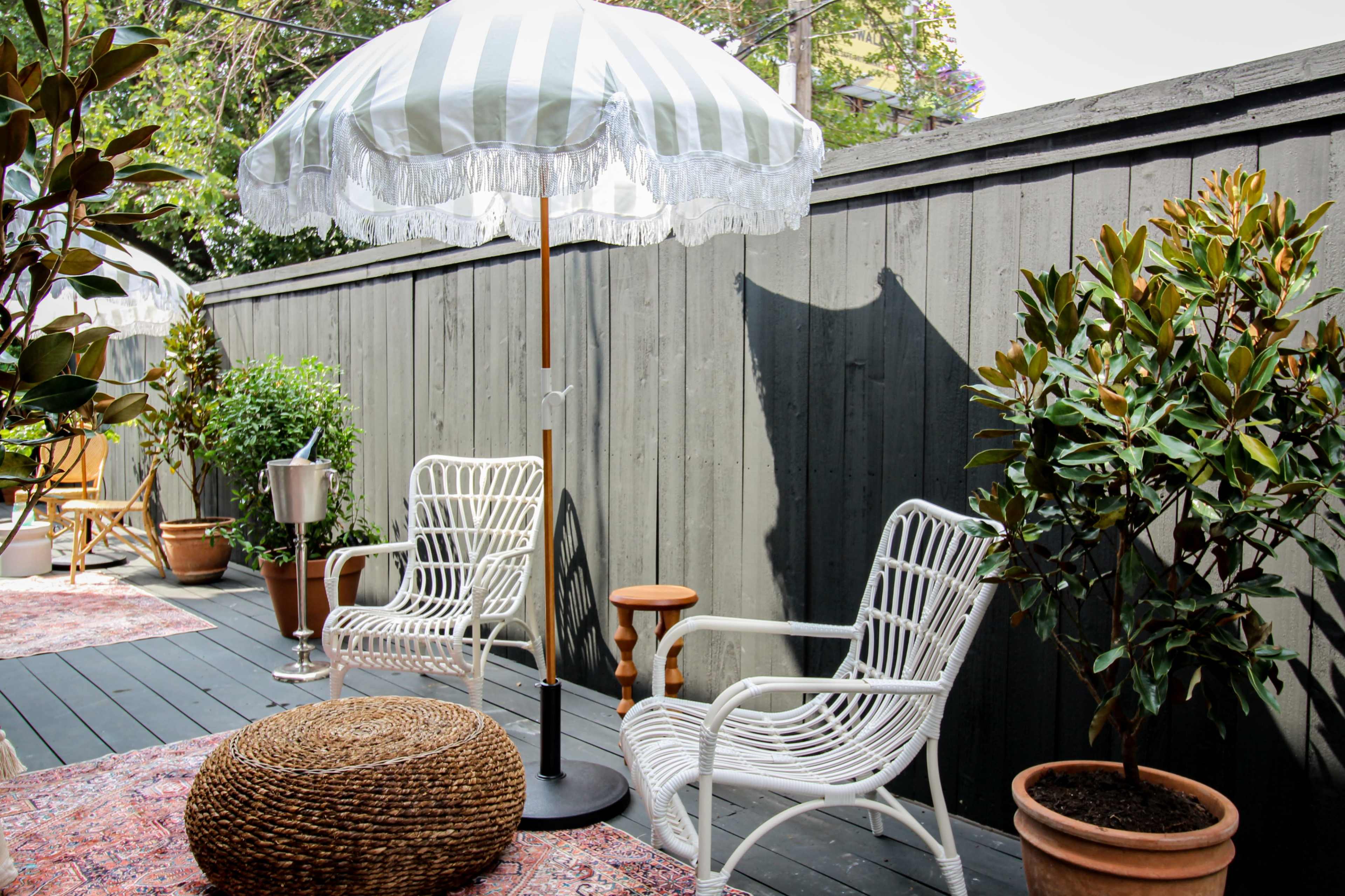 A patio area features two white wicker chairs under a striped umbrella, surrounded by potted plants and a decorative pouf on a wooden deck.