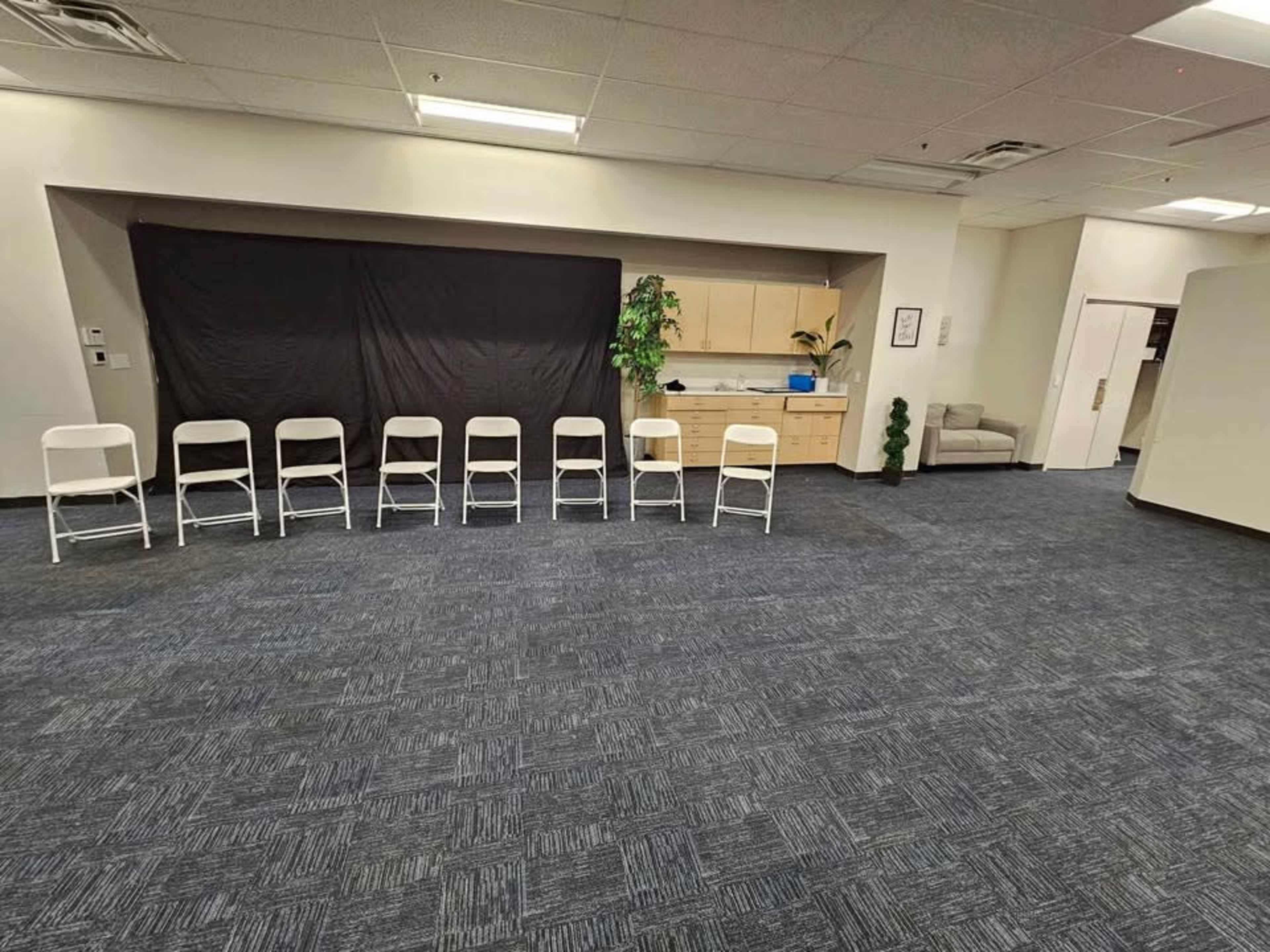 The image shows a large, empty room with a row of white chairs positioned in front of a black curtain, along with a few plants and a kitchenette area in the background.