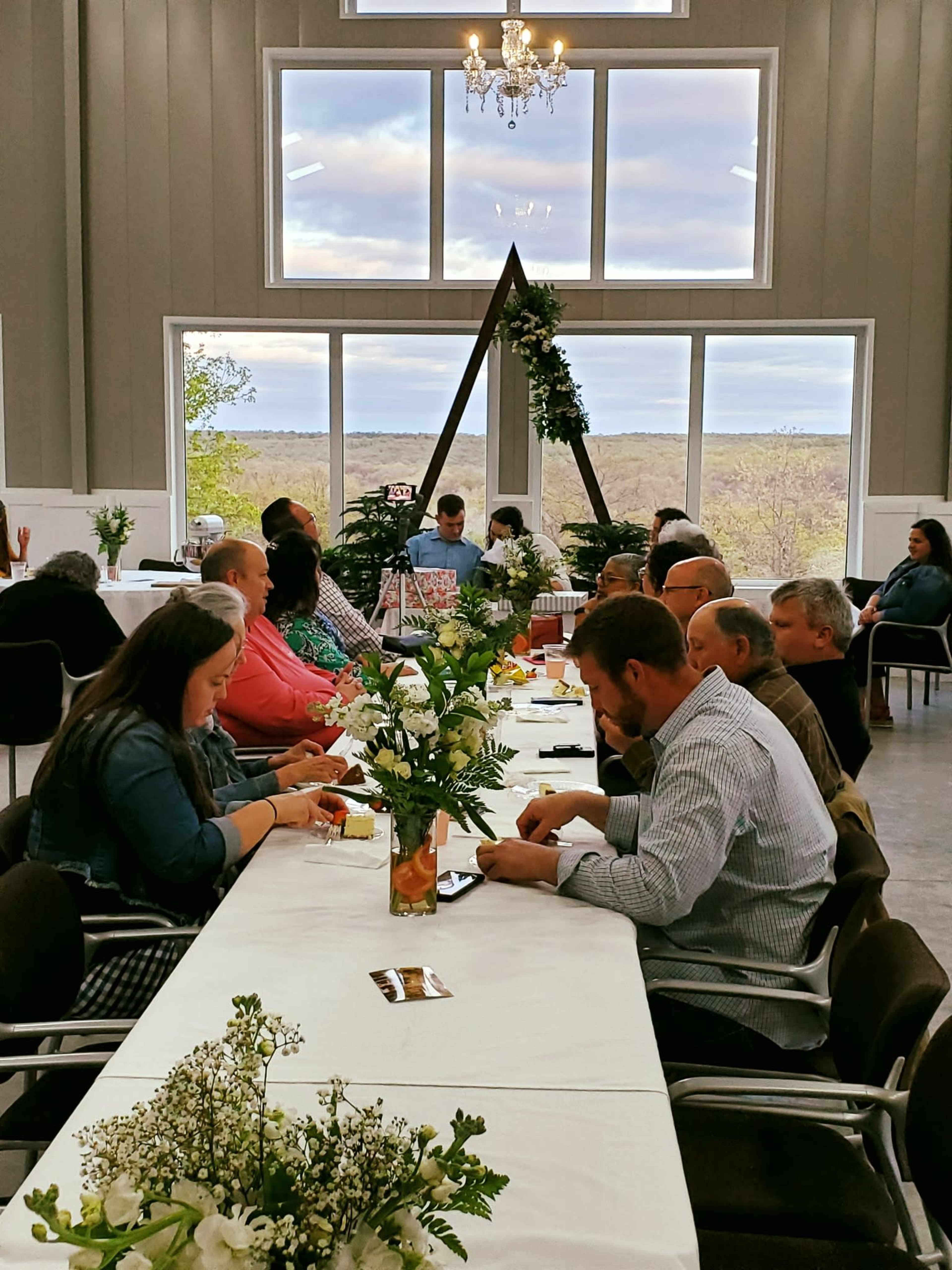 A long table is set for a gathering in a brightly lit room with large windows overlooking a landscape, with guests seated on either side.