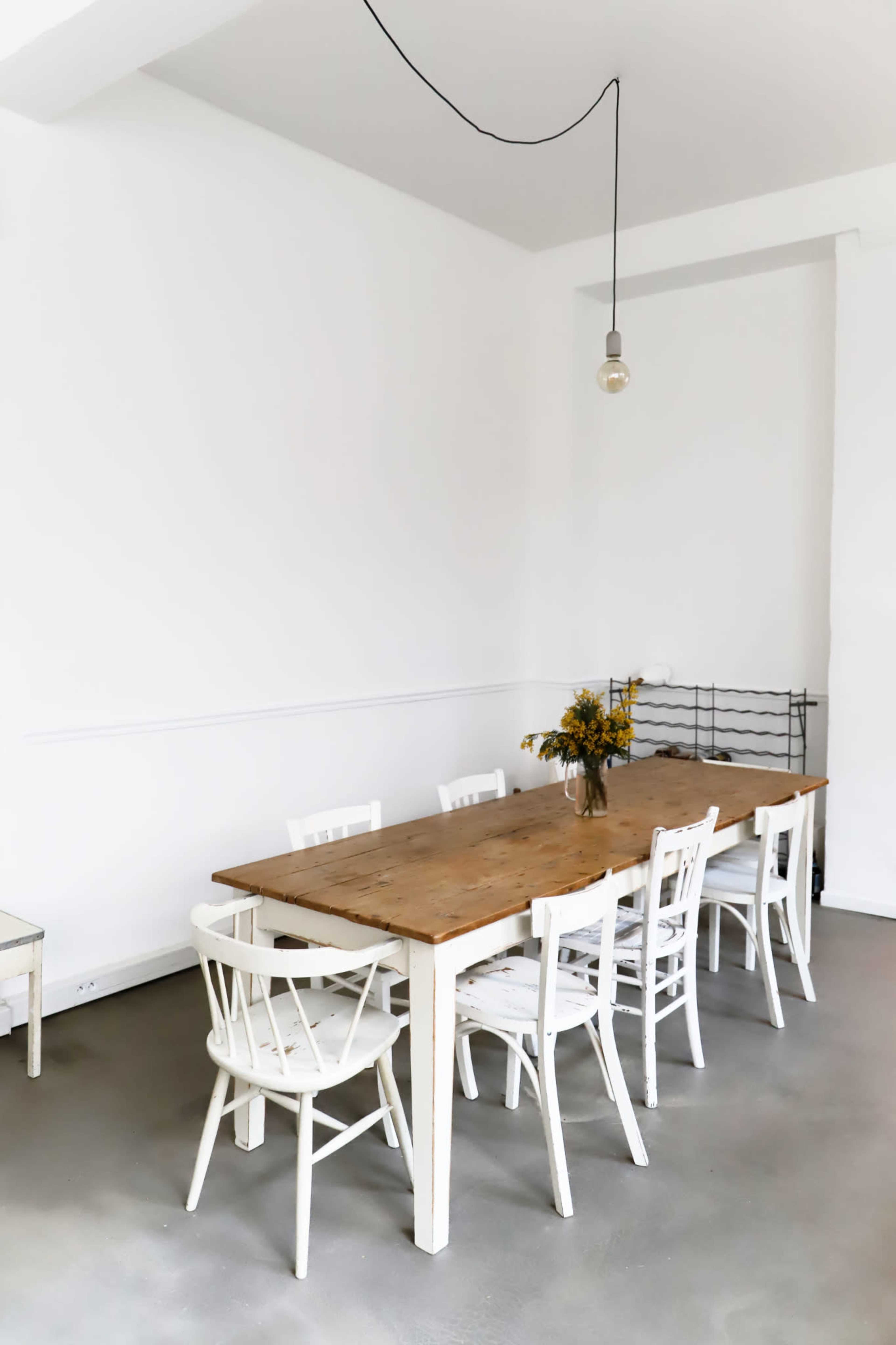 A minimalistic dining area features a long wooden table surrounded by white chairs and a vase of flowers.