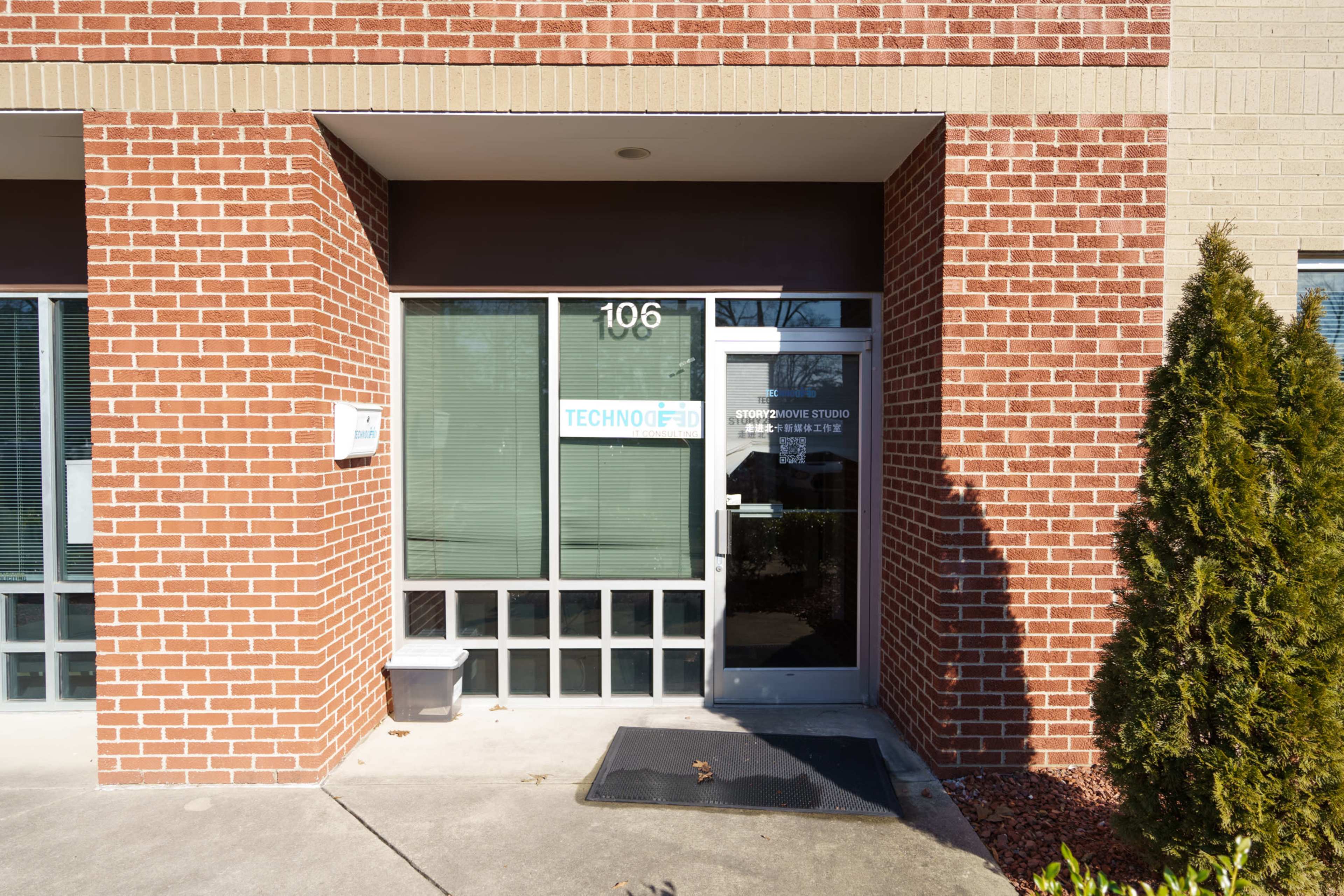 The entrance of a brick building with a glass door labeled "TECHNOFIT" and window shades, framed by a small bush and a utility box nearby.
