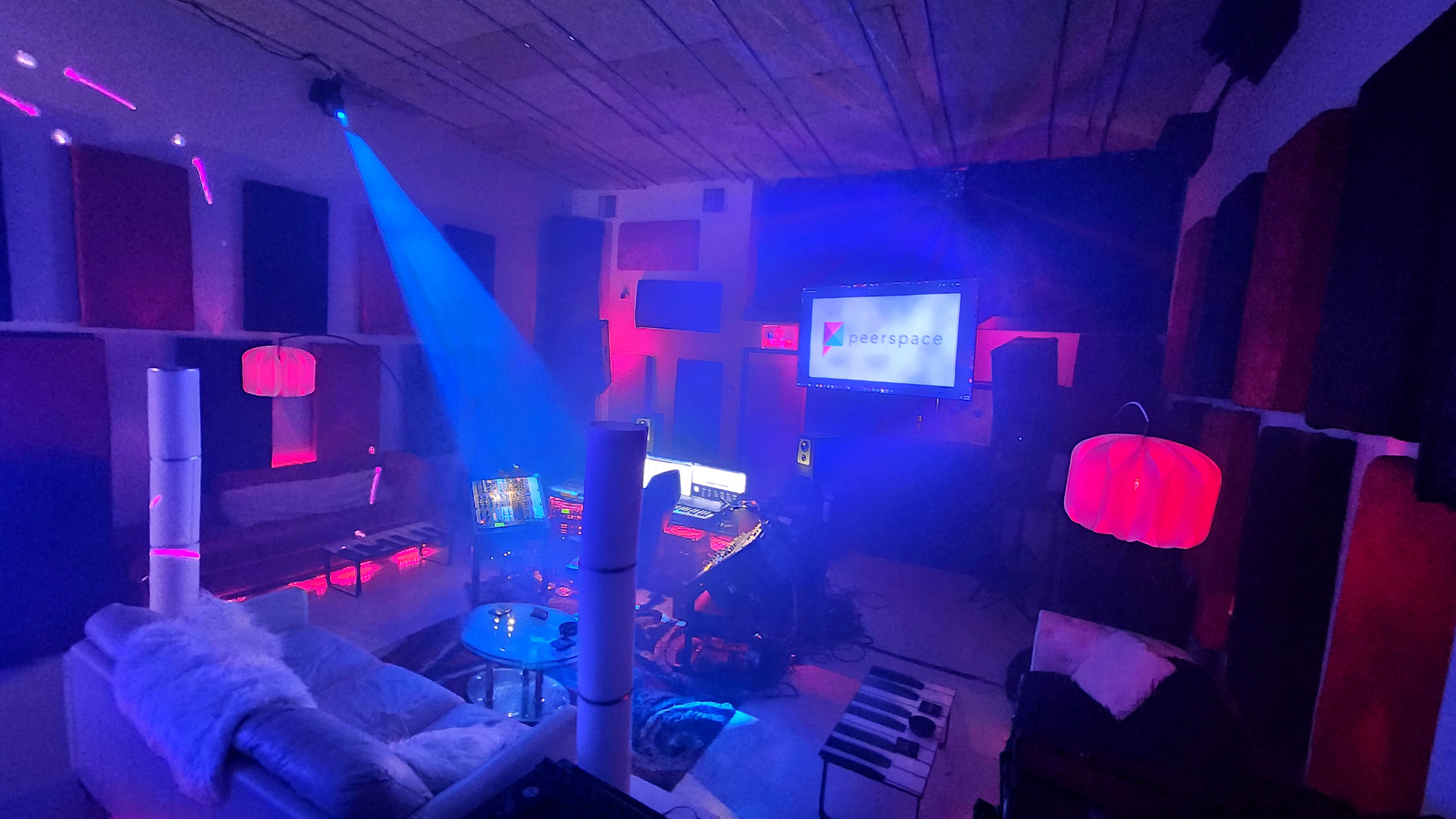 A dimly lit music studio with colorful LED lighting, a computer setup, and soundproofing panels on the walls.