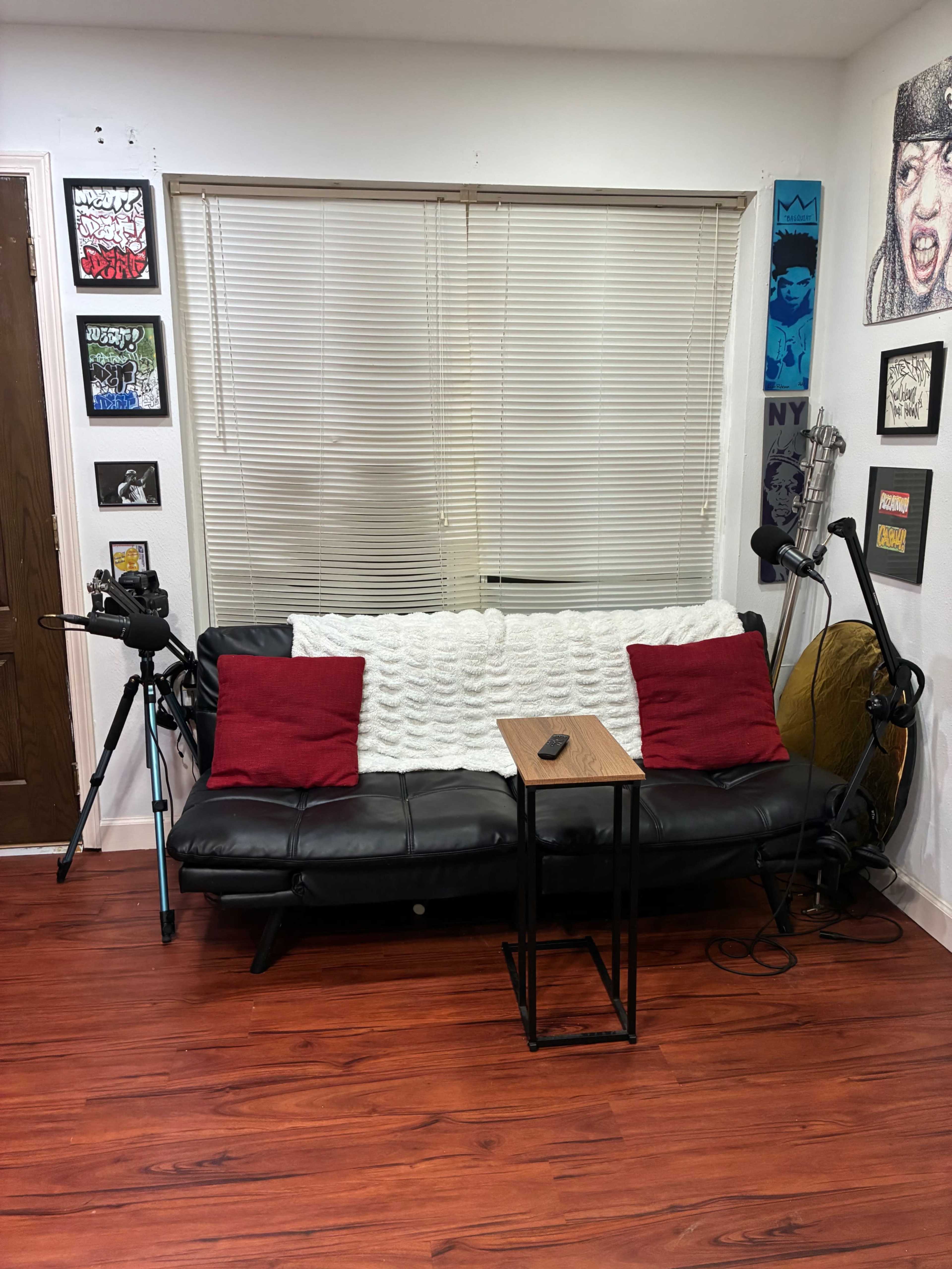 The image shows a small sitting area with a black couch adorned with red cushions, a wooden table, and a camera on a tripod, along with various framed artworks on the walls.