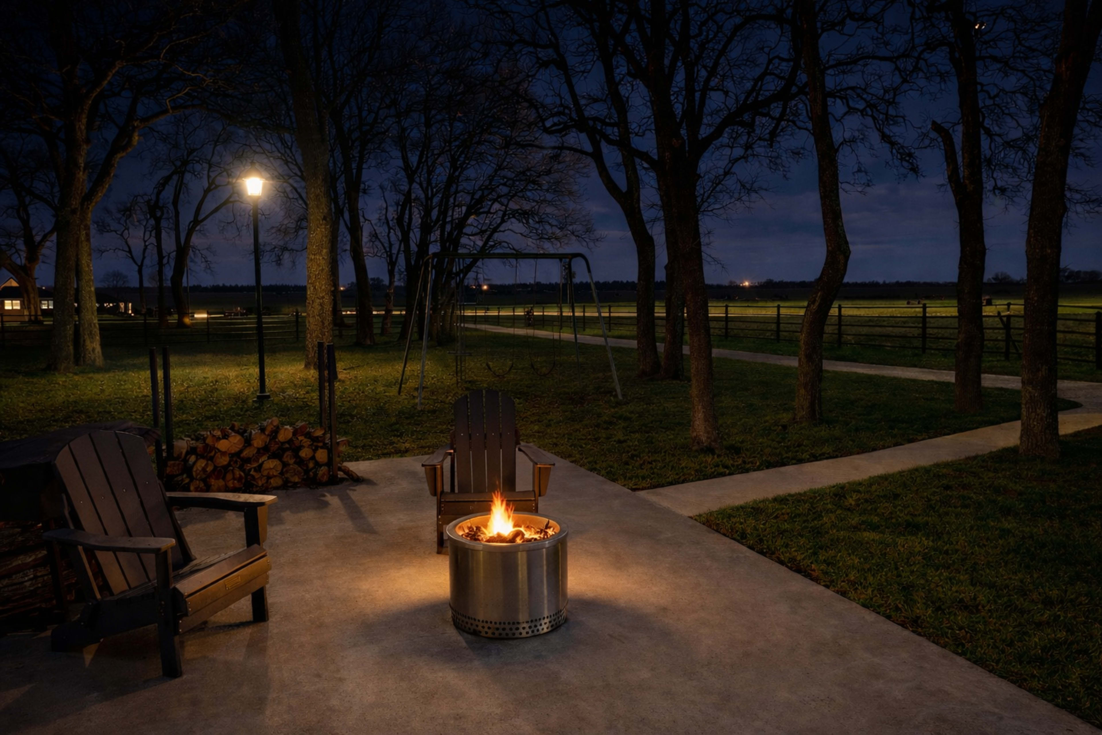 A fire pit surrounded by two wooden chairs sits on a concrete patio, illuminated by a lamp in a dark, wooded area.