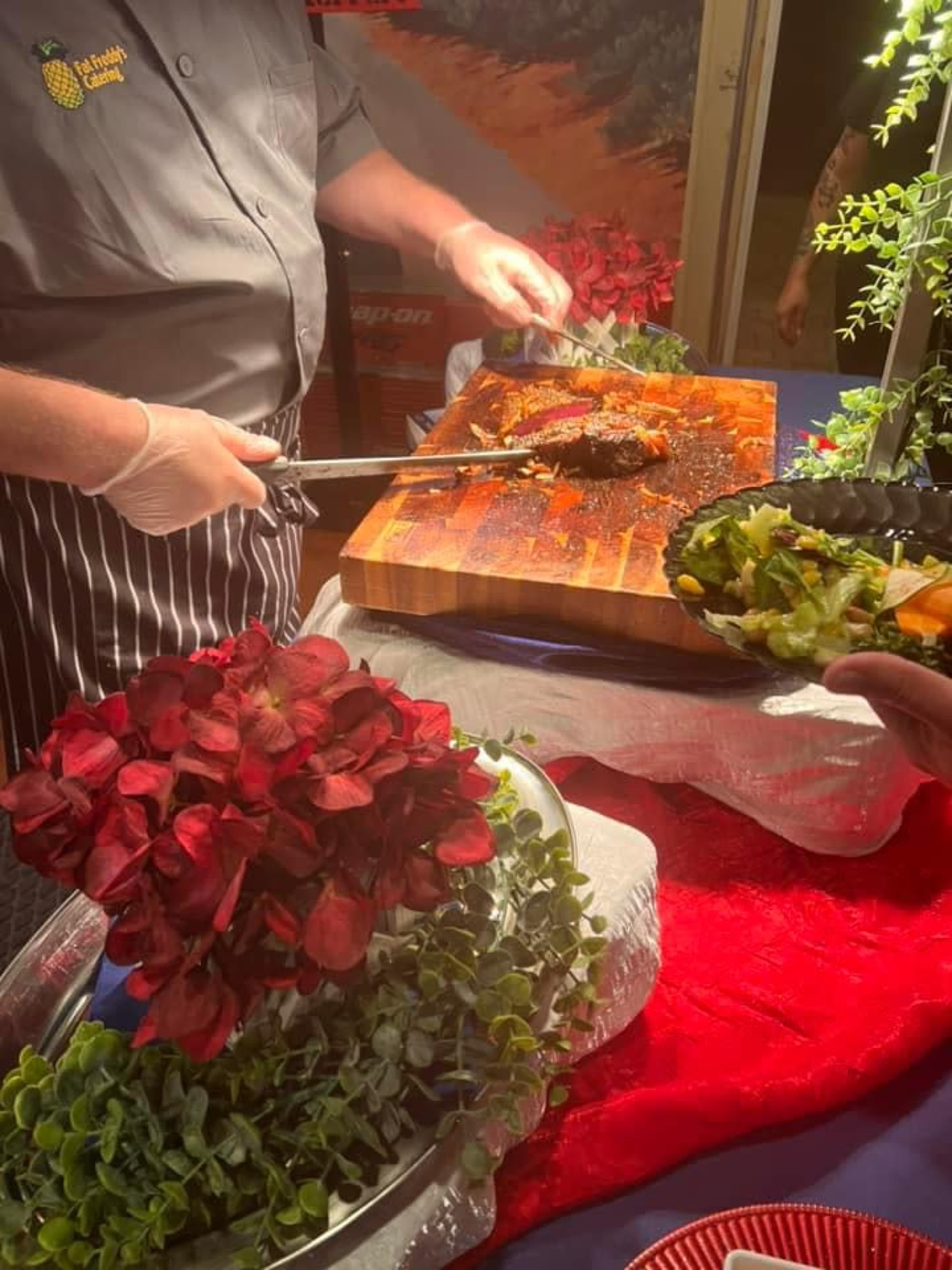 A chef carves a piece of meat on a wooden cutting board, surrounded by colorful decorations and a hand holding a salad bowl.