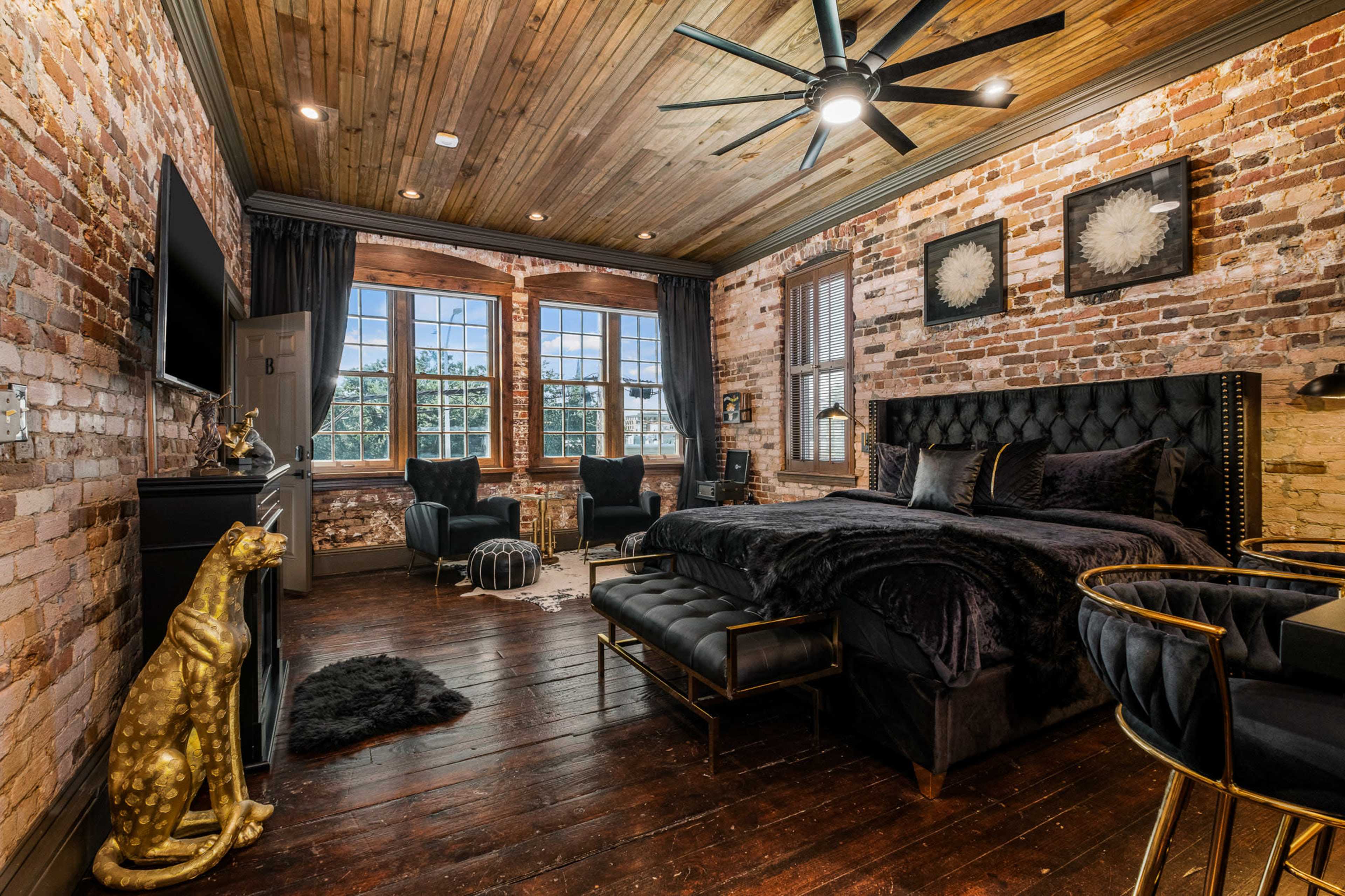 A cozy bedroom featuring exposed brick walls, a wooden ceiling, a king-size bed with dark bedding, and two armchairs near large windows.