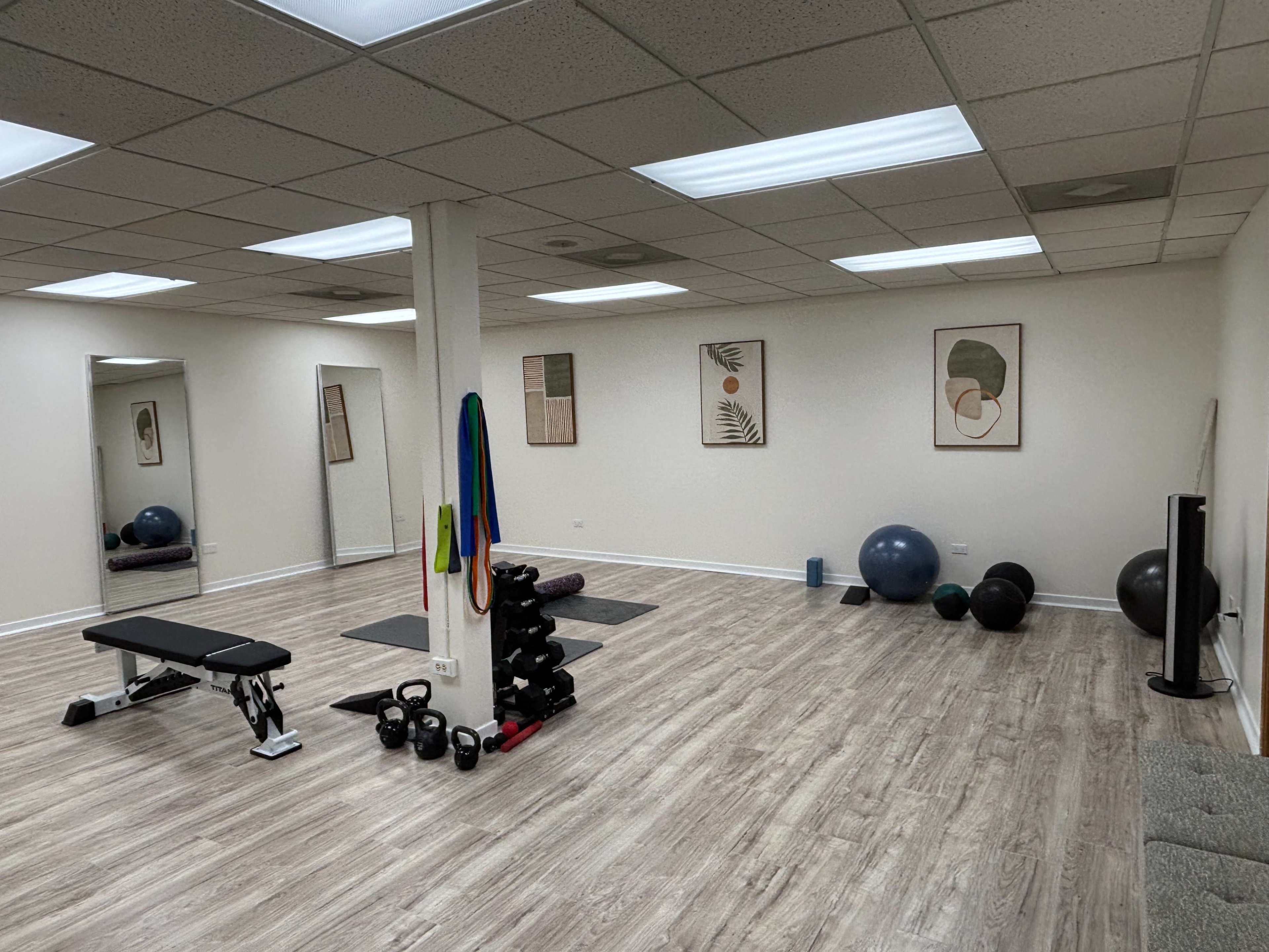 The image shows a spacious fitness studio equipped with a bench, free weights, exercise balls, and resistance bands, along with a large mirror on one wall.
