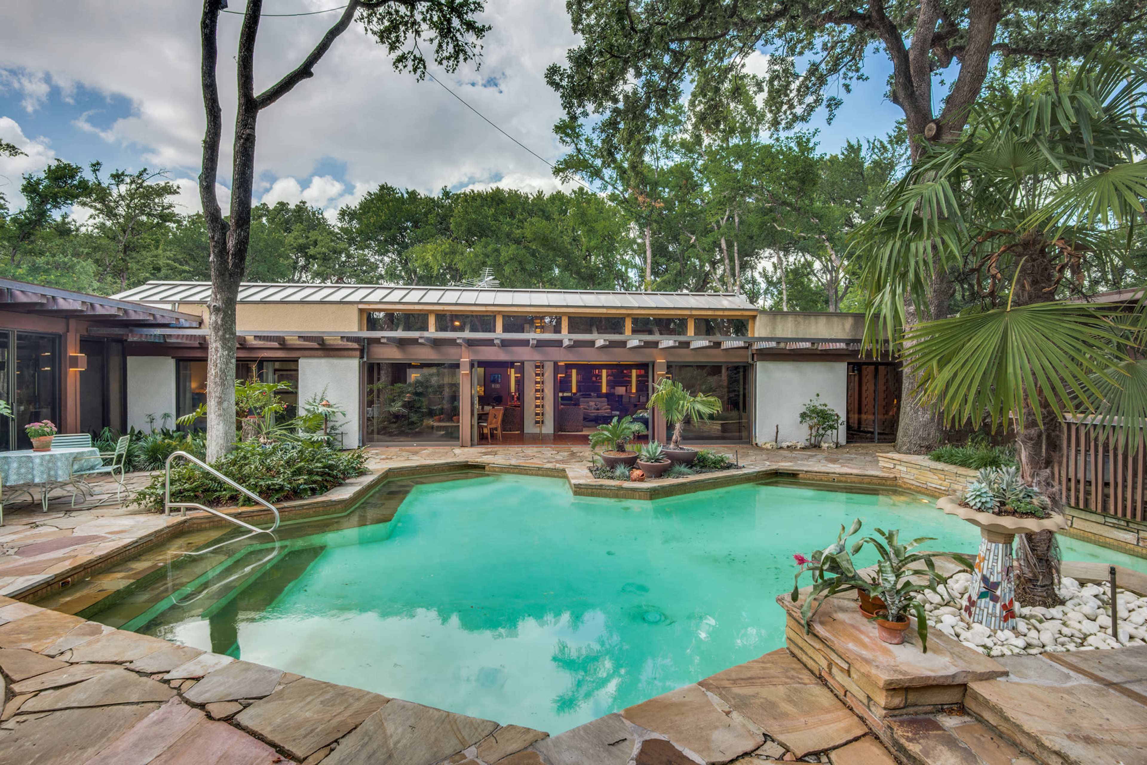 A backyard with a green pool surrounded by stone decking, tropical plants, and a house in the background with large windows.